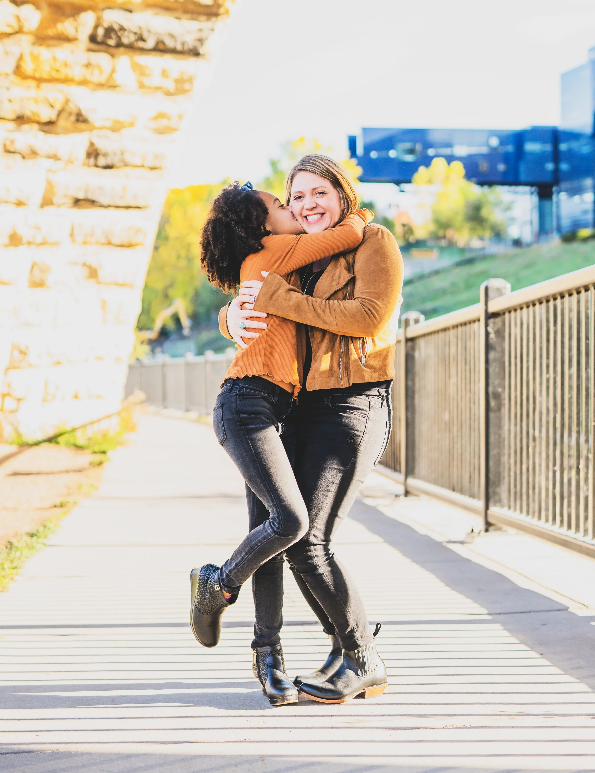 A young girl hugging and kissing a smiling woman outdoors on a sunny day, with a bridge and modern buildings in the background.