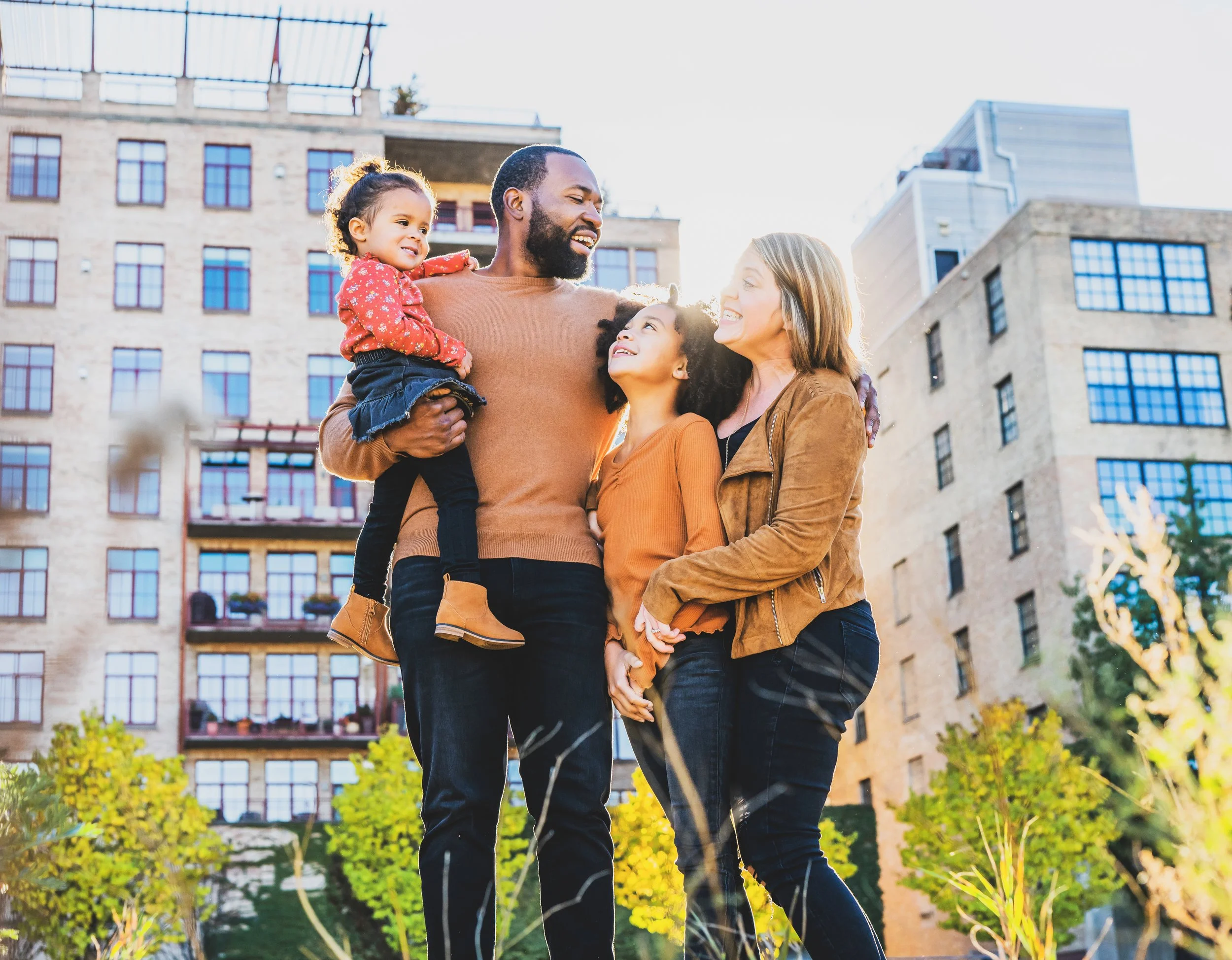 A diverse family of four smiling and enjoying time together outdoors in an urban park with high-rise buildings behind them.