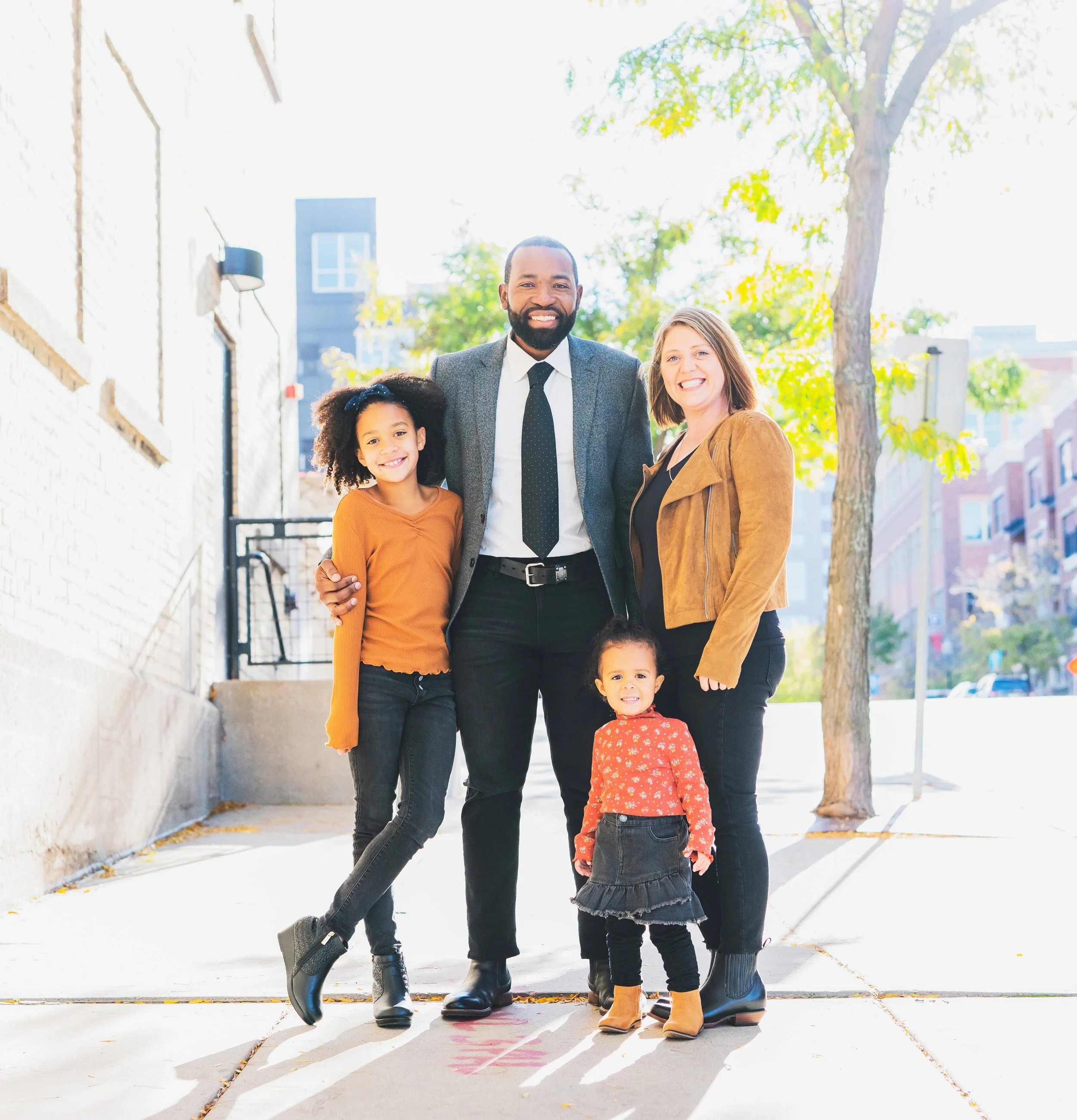 A happy multiracial family of four standing together on a sidewalk in an urban neighborhood during daytime, smiling at the camera.