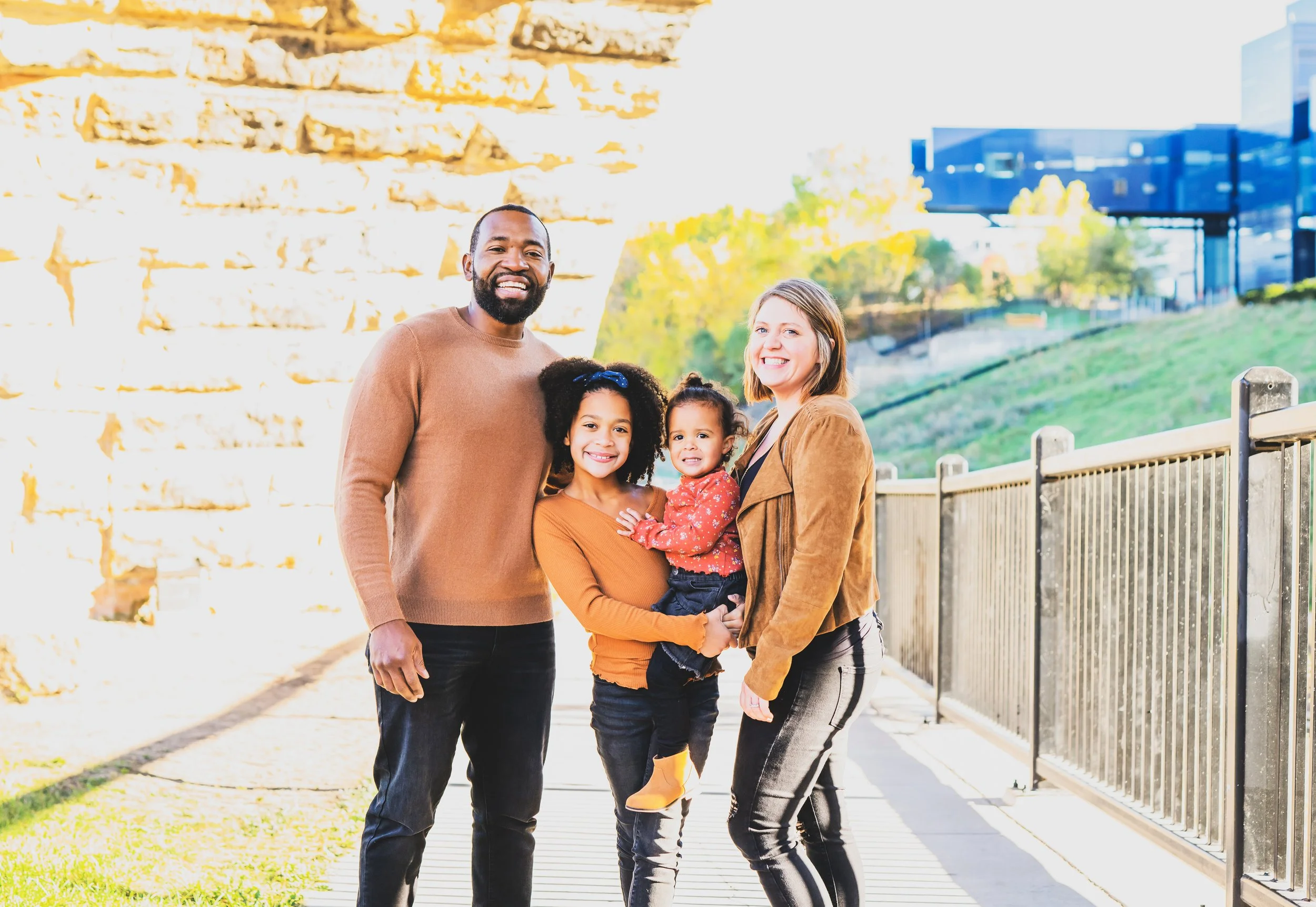 A diverse family of four, including a man, a woman, and two young girls, stands together outdoors on a sunny day, smiling at the camera with a cityscape and greenery in the background.