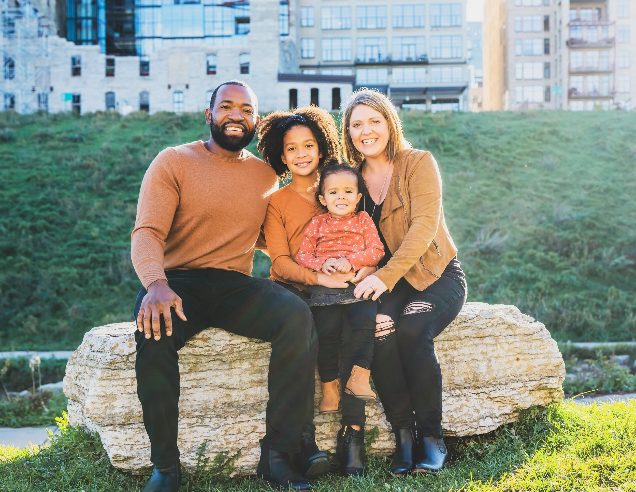 A family of four sitting together on a large rock outdoors with a cityscape background, smiling at the camera