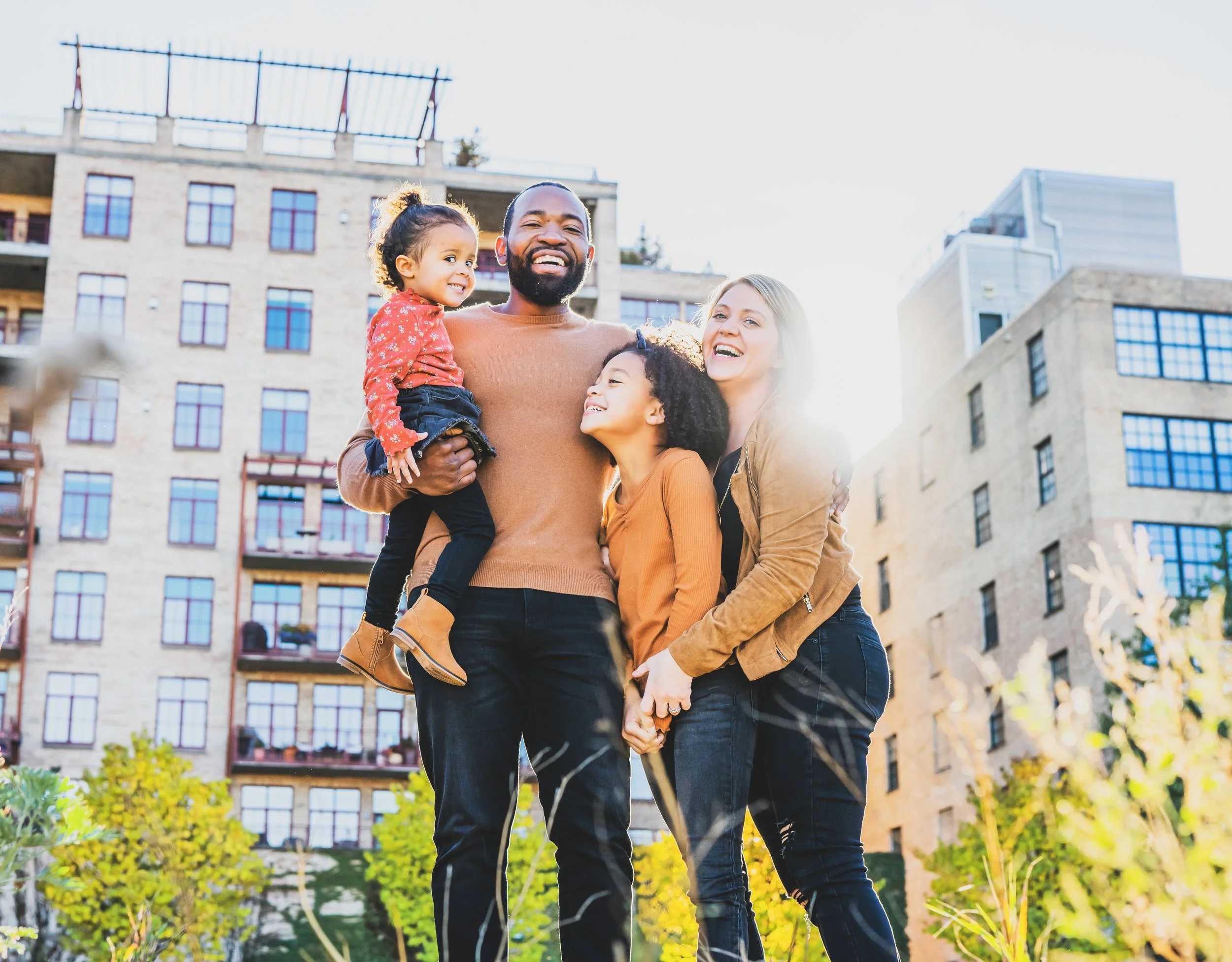A happy family of four, including two women, one man, and two young girls, enjoying outdoors in an urban park with apartment buildings in the background, during daytime.