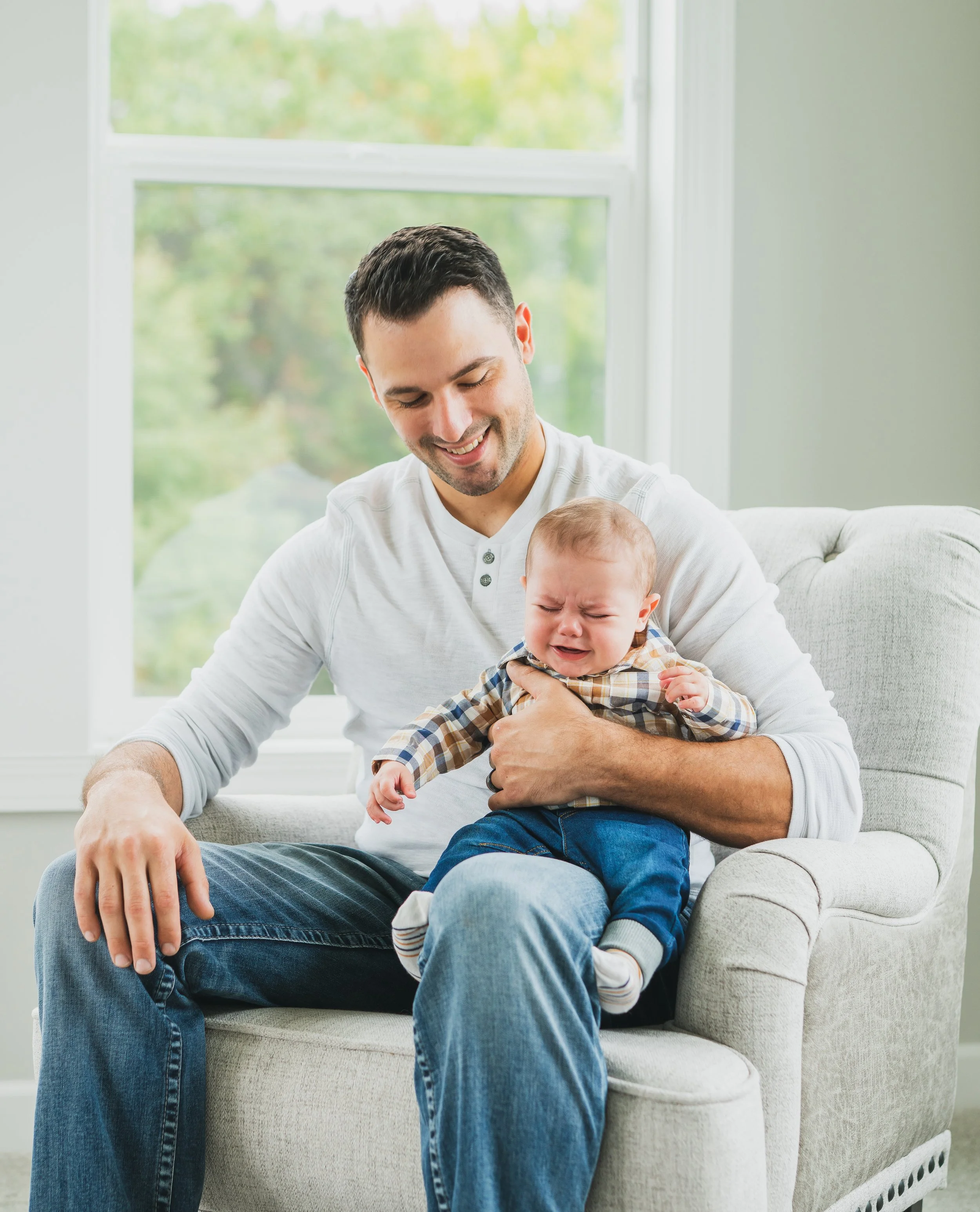 A man smiling at a crying baby sitting on his lap on a beige armchair in a sunny room with large windows.