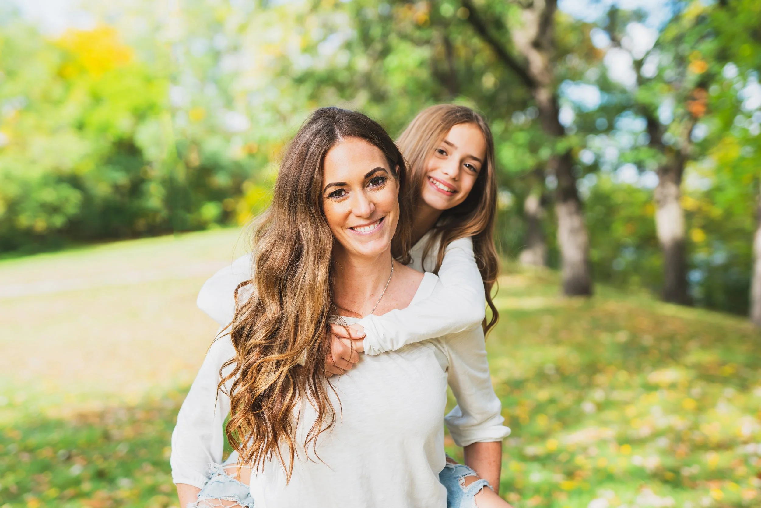 A woman giving a piggyback ride to a young girl in a park with trees and fall foliage.