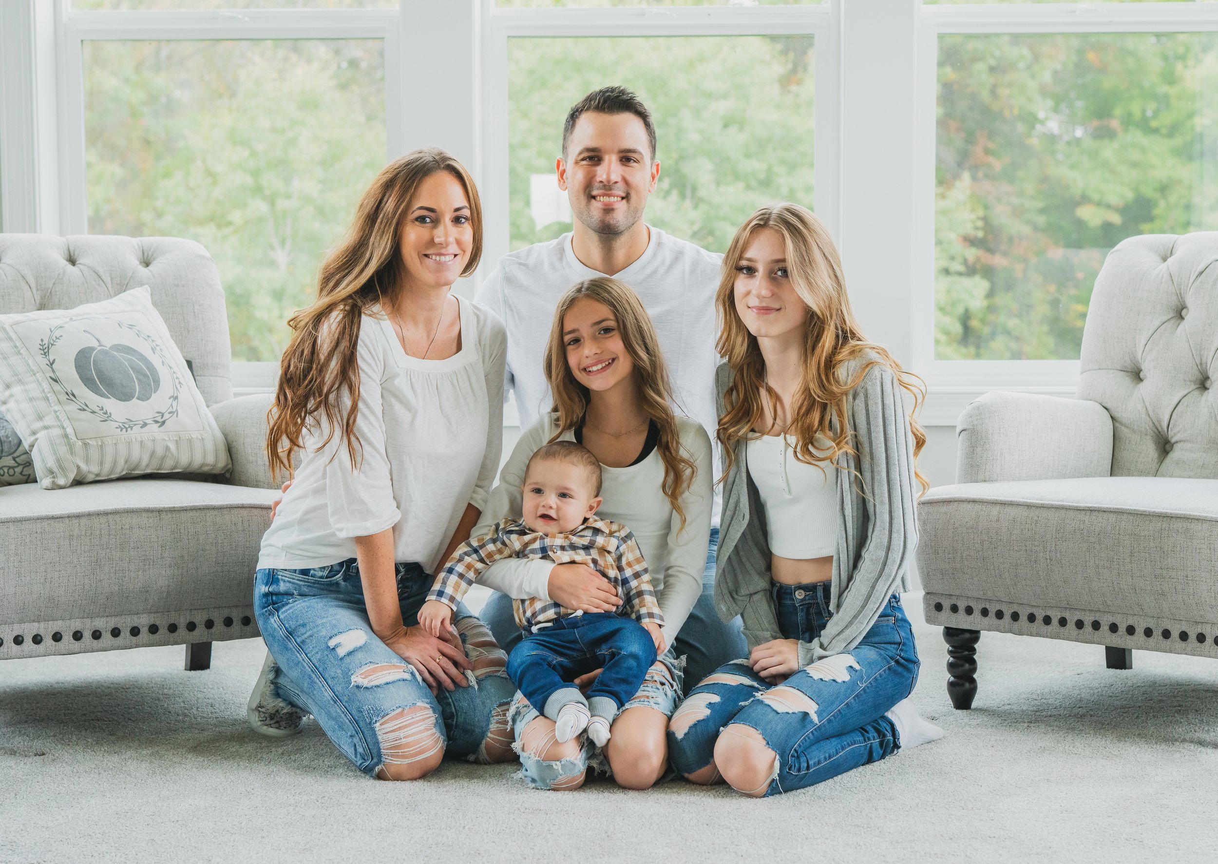 Family portrait of six people, including three women, one man, and two children, sitting on a light-colored carpet in a bright living room with large windows and green trees outside.