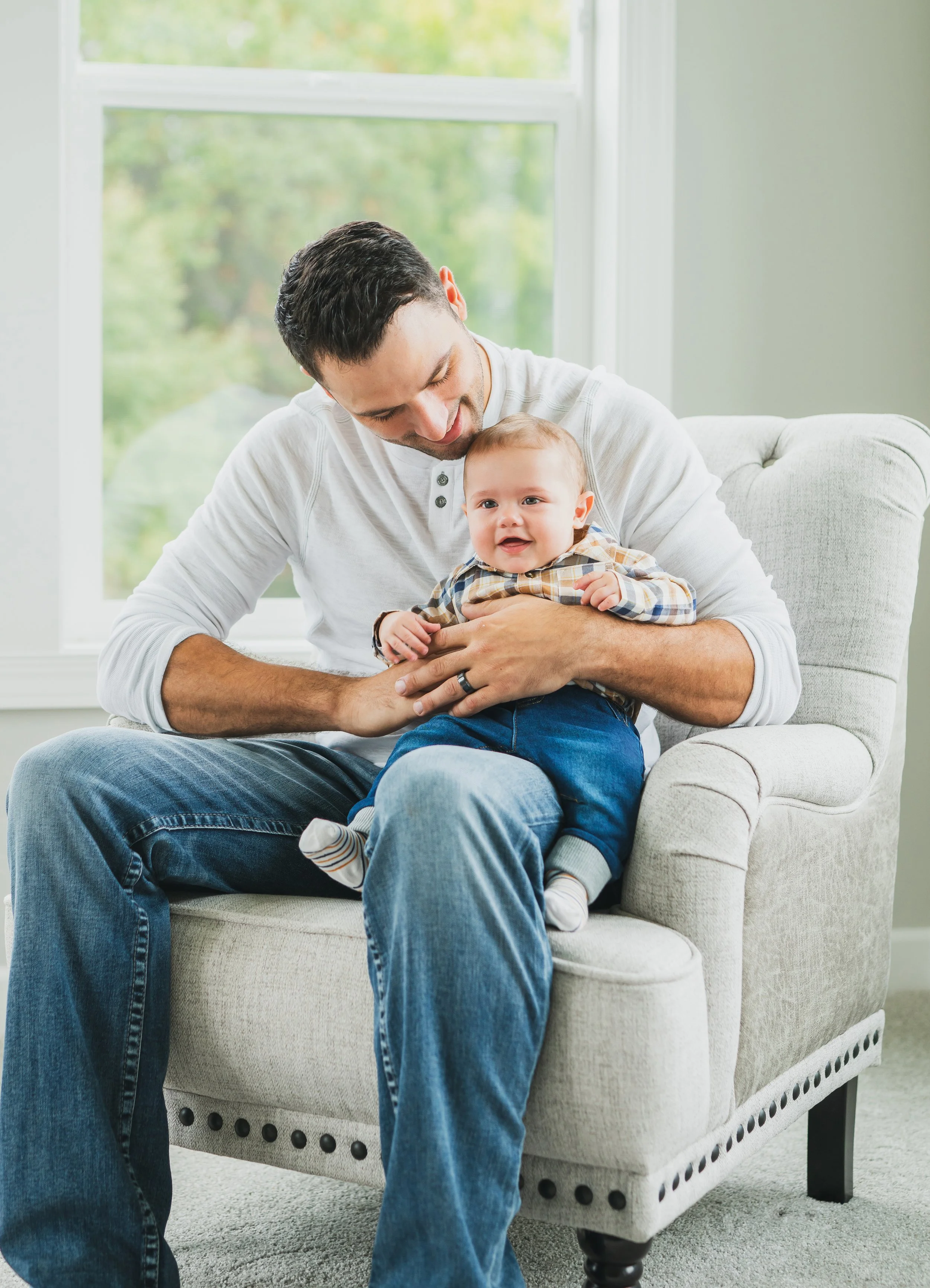 A man holding a smiling baby girl on his lap while sitting in a beige armchair in a room with large windows and green outdoor scenery.