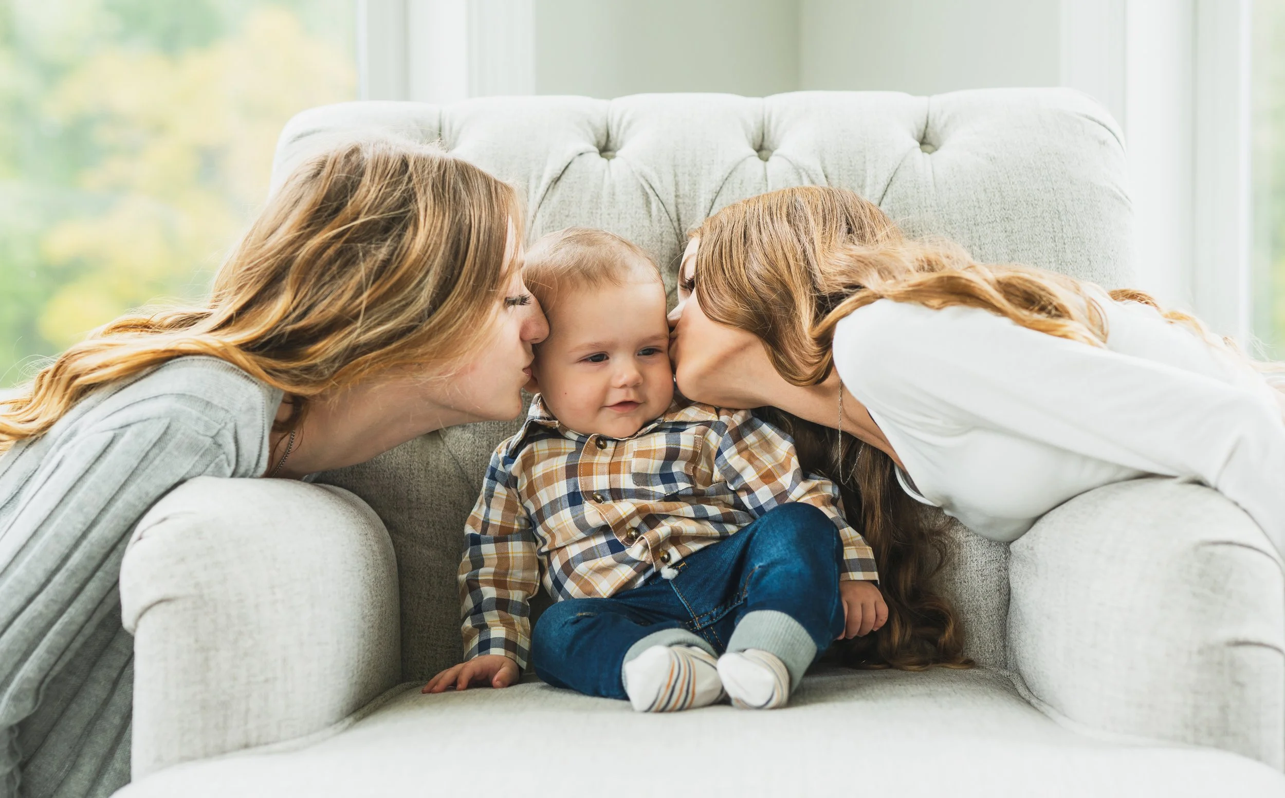 Two women kissing a toddler on the cheeks, sitting on a light-colored couch in a bright room.