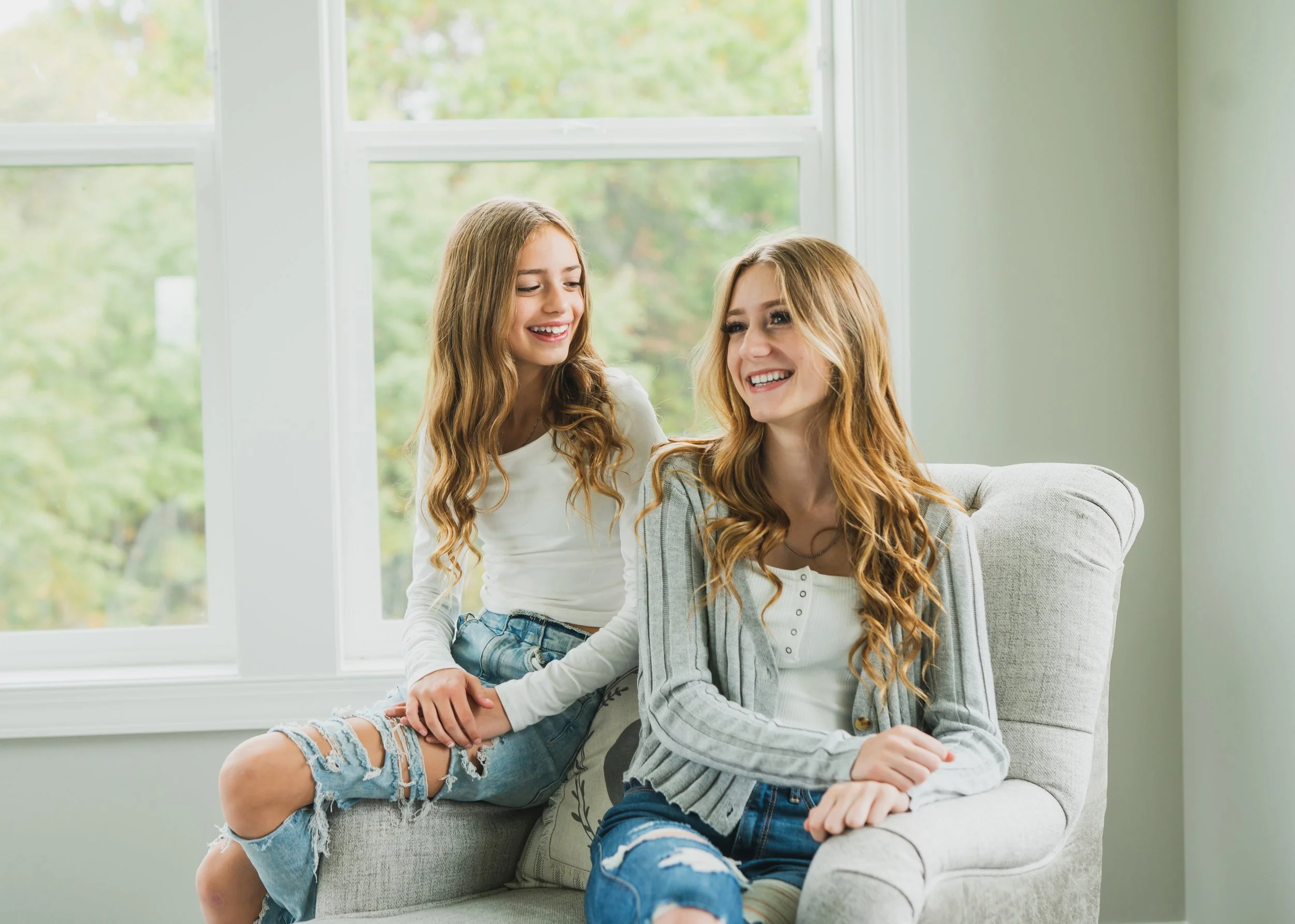 Two young women sitting on a light-colored armchair by a large window with green trees outside, smiling and enjoying conversation.