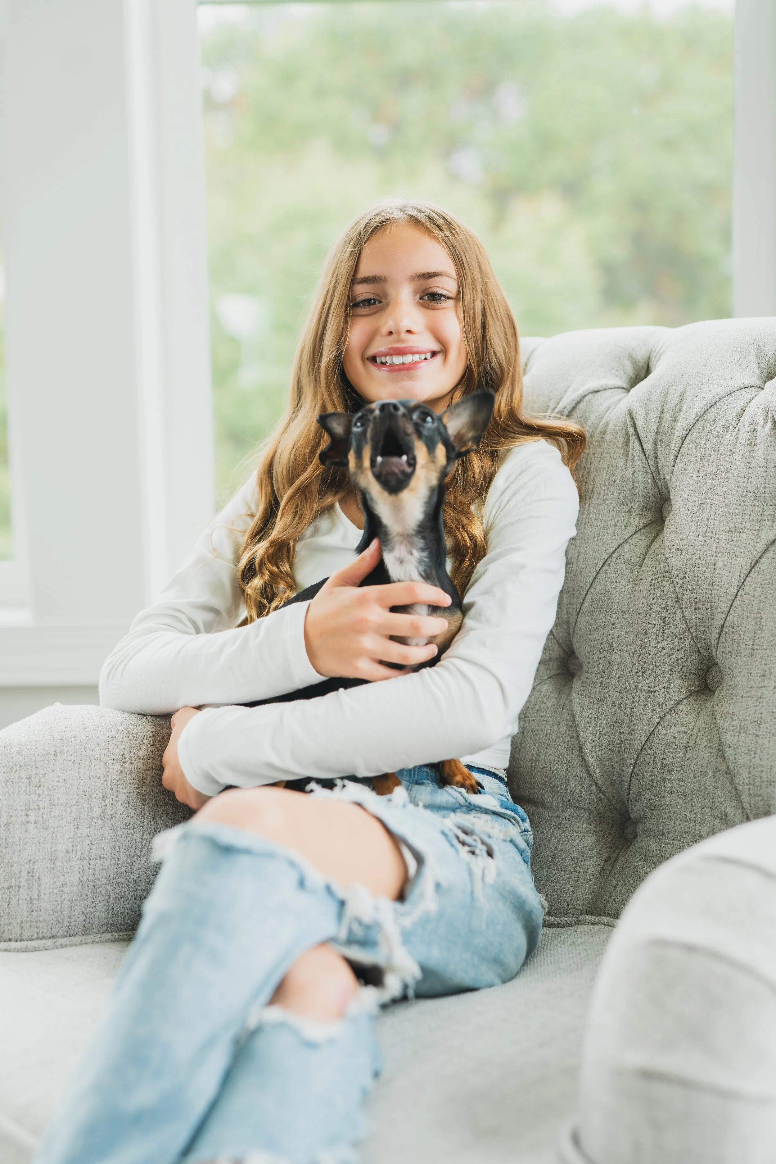 A young woman with long wavy hair sitting on a light gray couch, holding a small black and tan Chihuahua dog, smiling at the camera inside a bright room with large windows showing green trees outside.