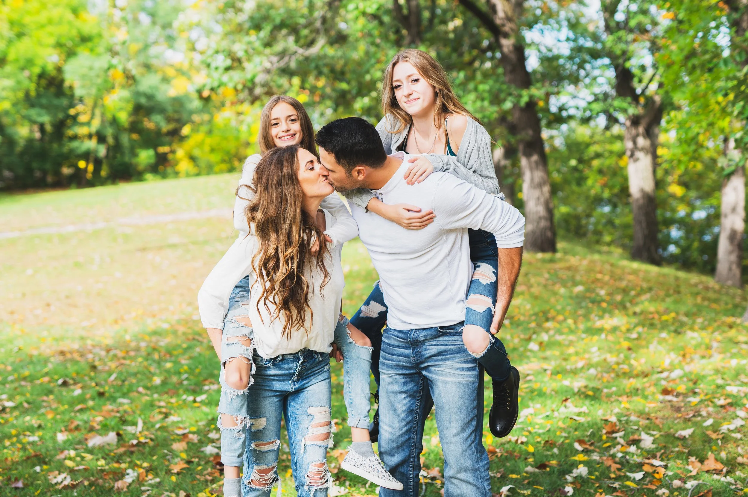 A family of four outdoors in a park, a woman and a man kissing, two young girls smiling nearby.