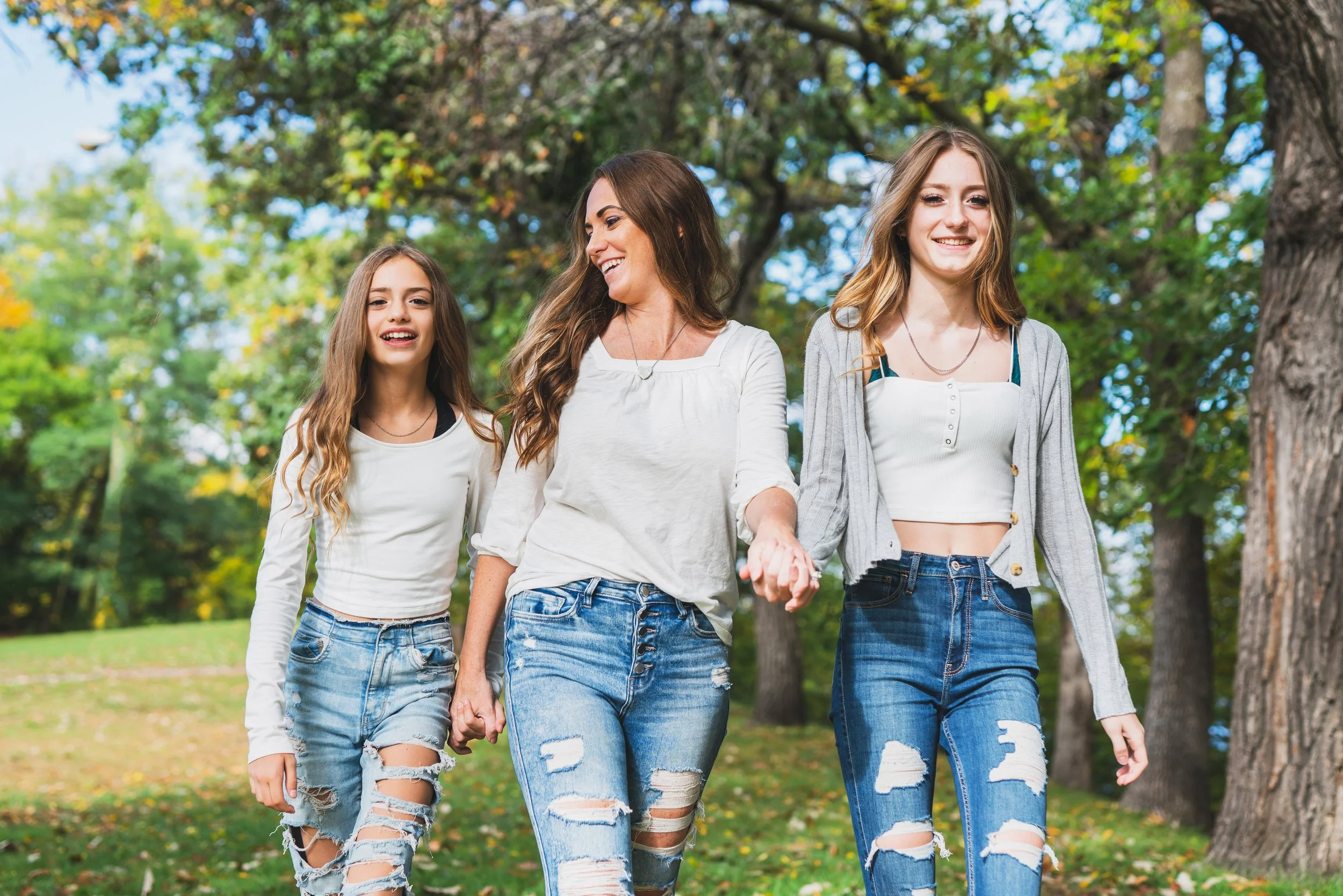 A woman and two teenage girls walking happily in a park, holding hands, surrounded by green trees and a grassy area.