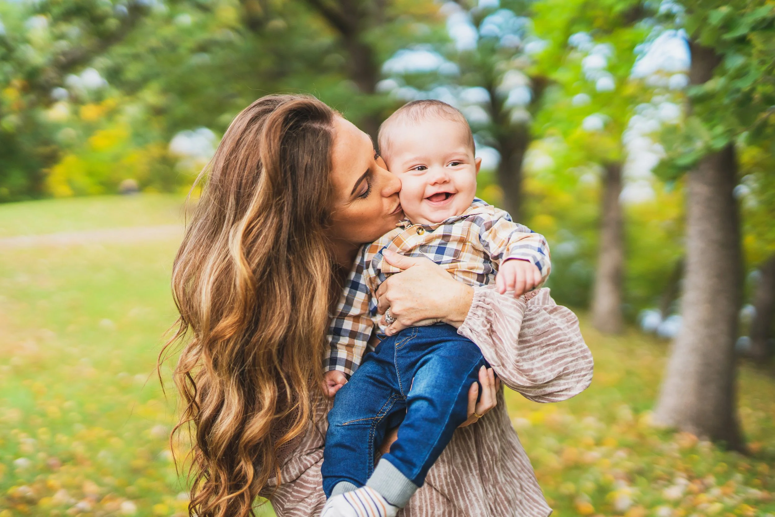 A woman with long, wavy brown hair holding a smiling baby boy outdoors in a park with trees.