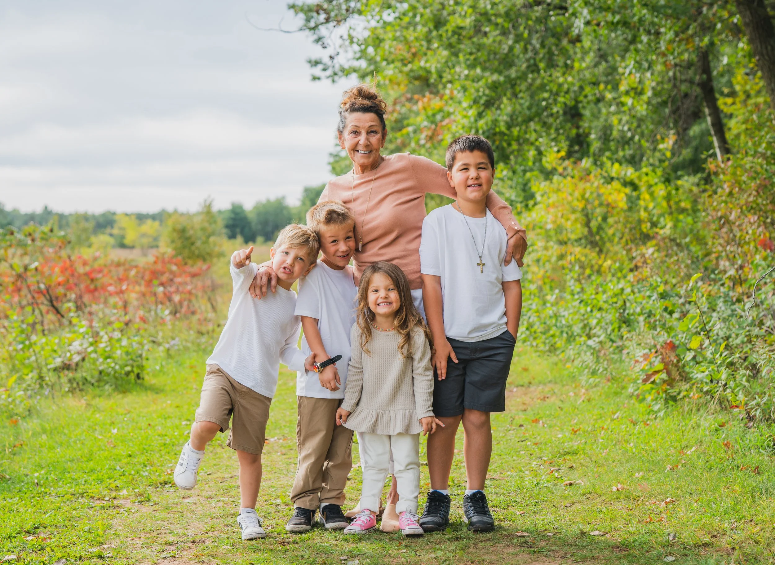 A smiling grandmother with four children walking on a nature trail in autumn, surrounded by trees with green and red leaves.