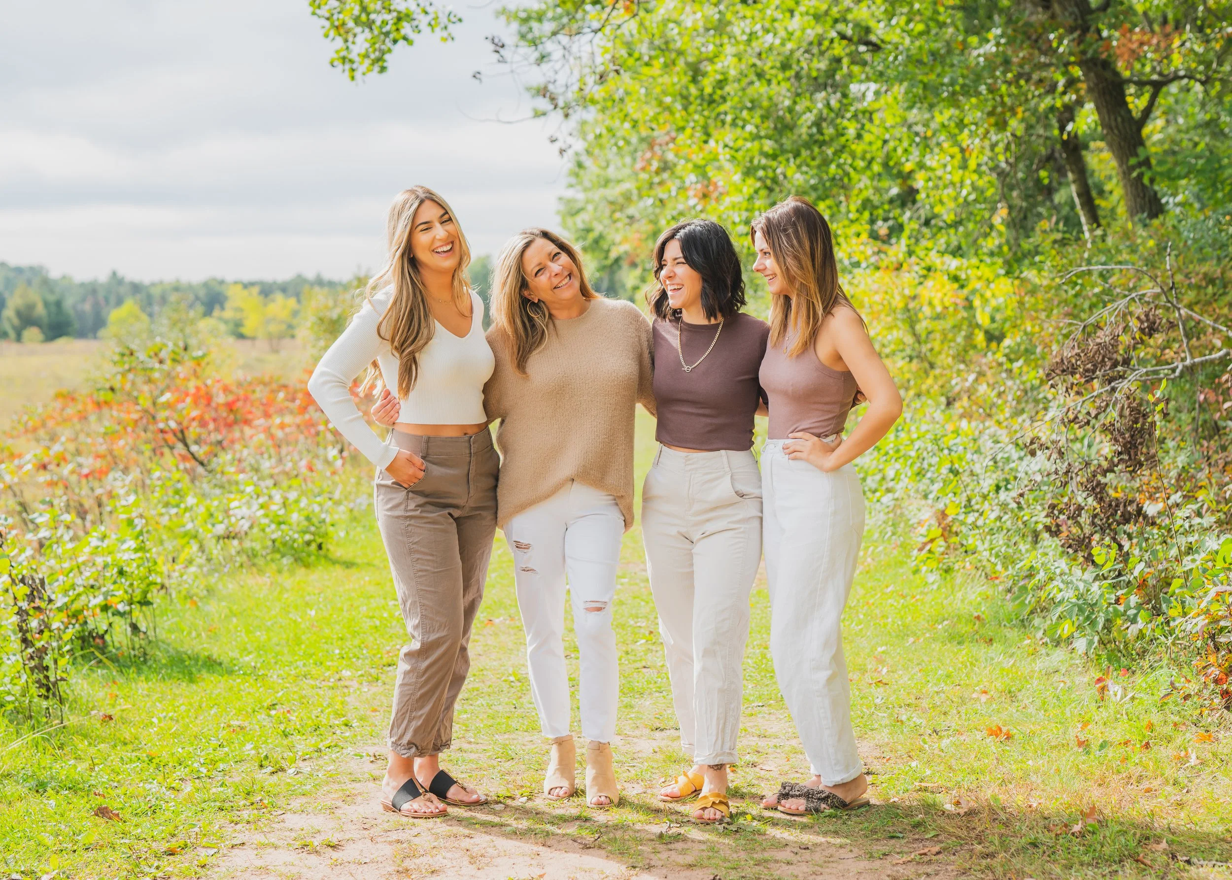 Four women standing close together on a dirt path in a lush, green outdoor setting, smiling and enjoying each other's company.