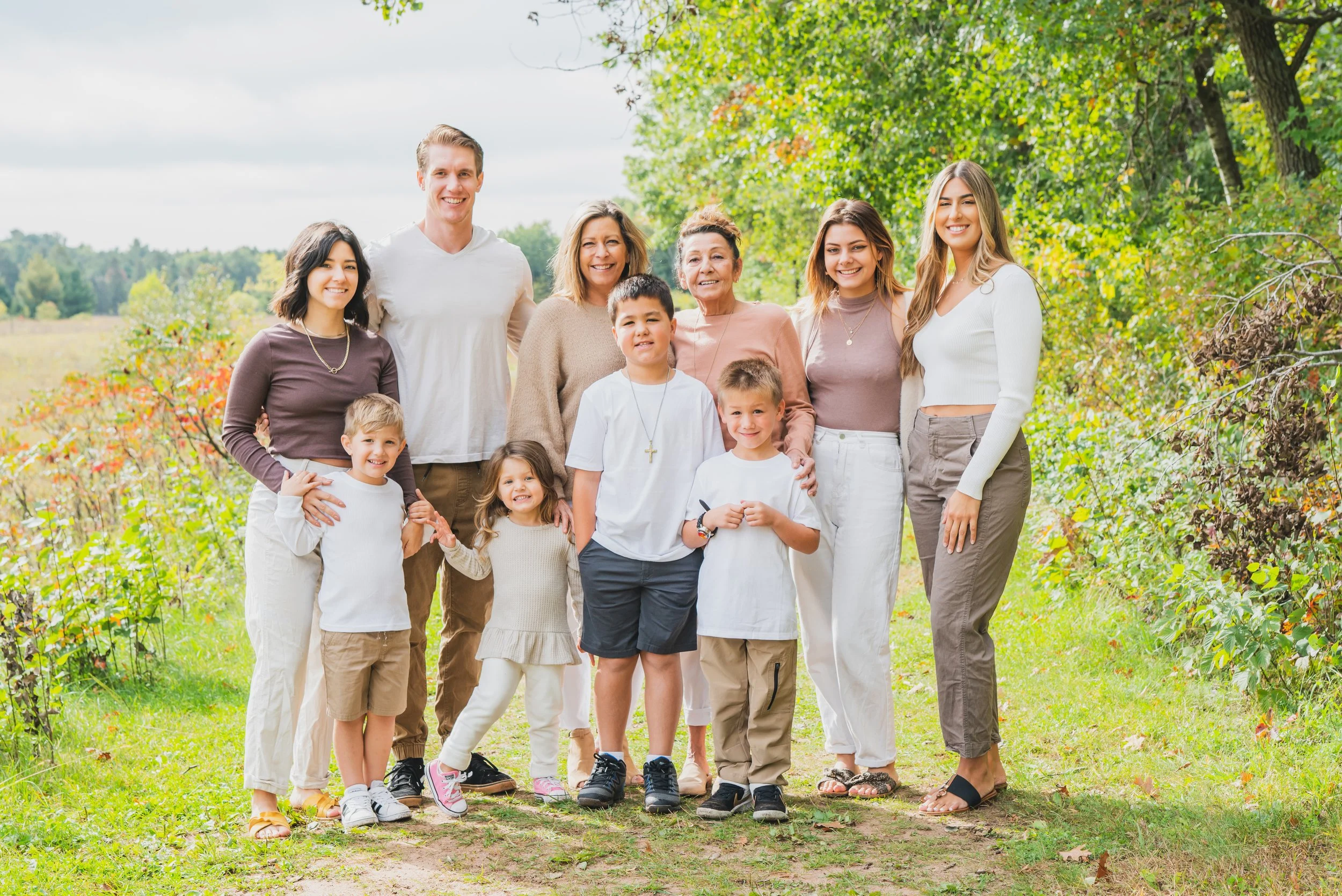 Family photo of three generations outdoors on a sunny day, standing on a grassy path surrounded by green trees and shrubs.