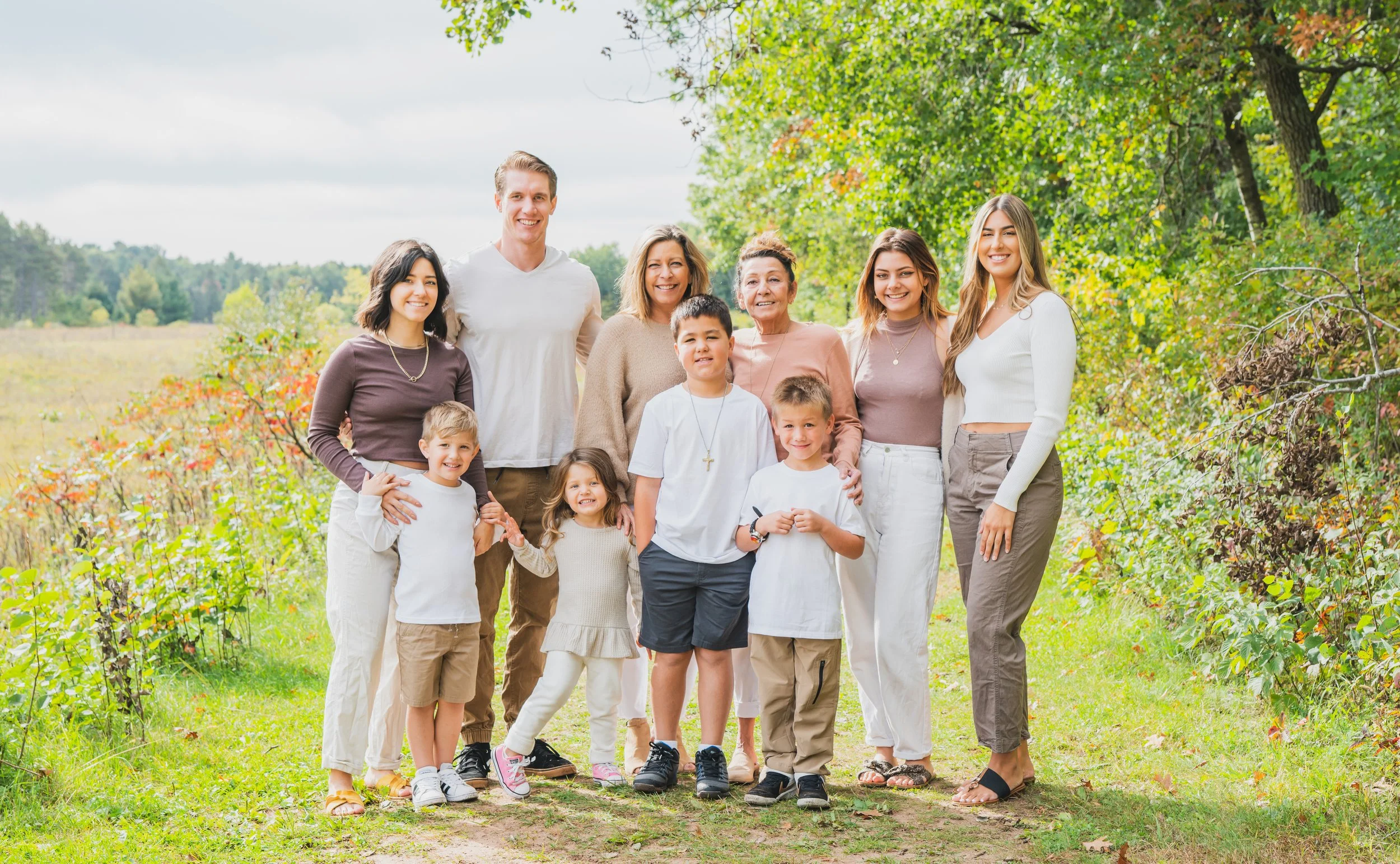 A multi-generational family group standing outdoors on a grassy path surrounded by trees, smiling at the camera.