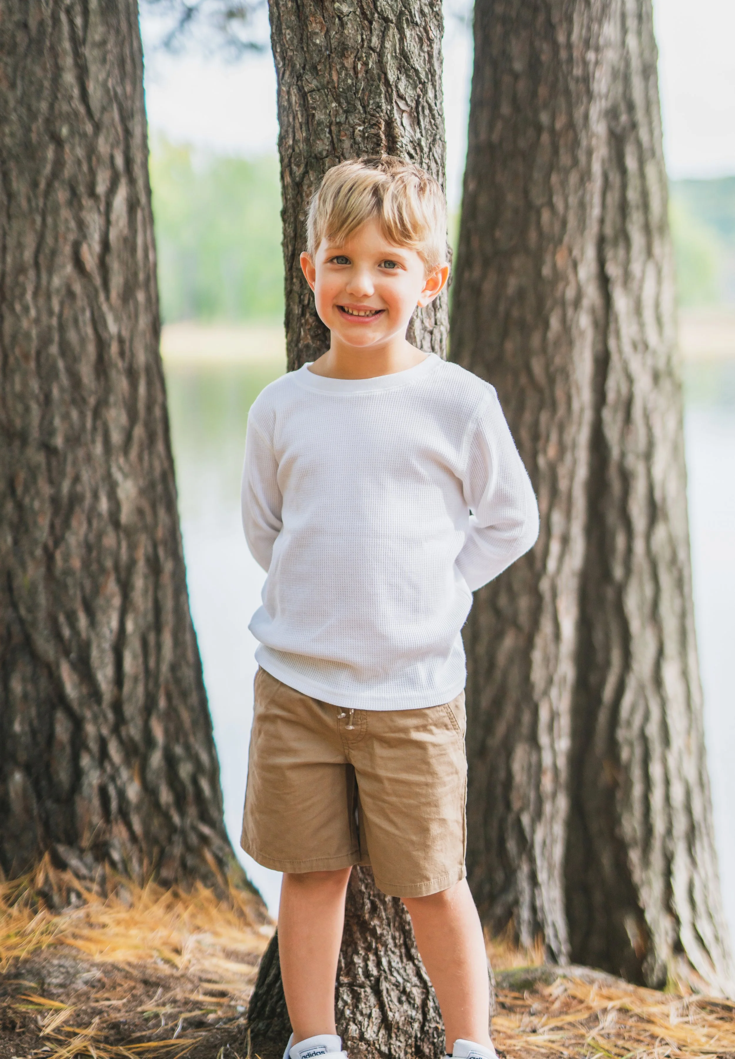 A young boy with blond hair smiling and standing outdoors in front of a tree, with a body of water and trees in the background.