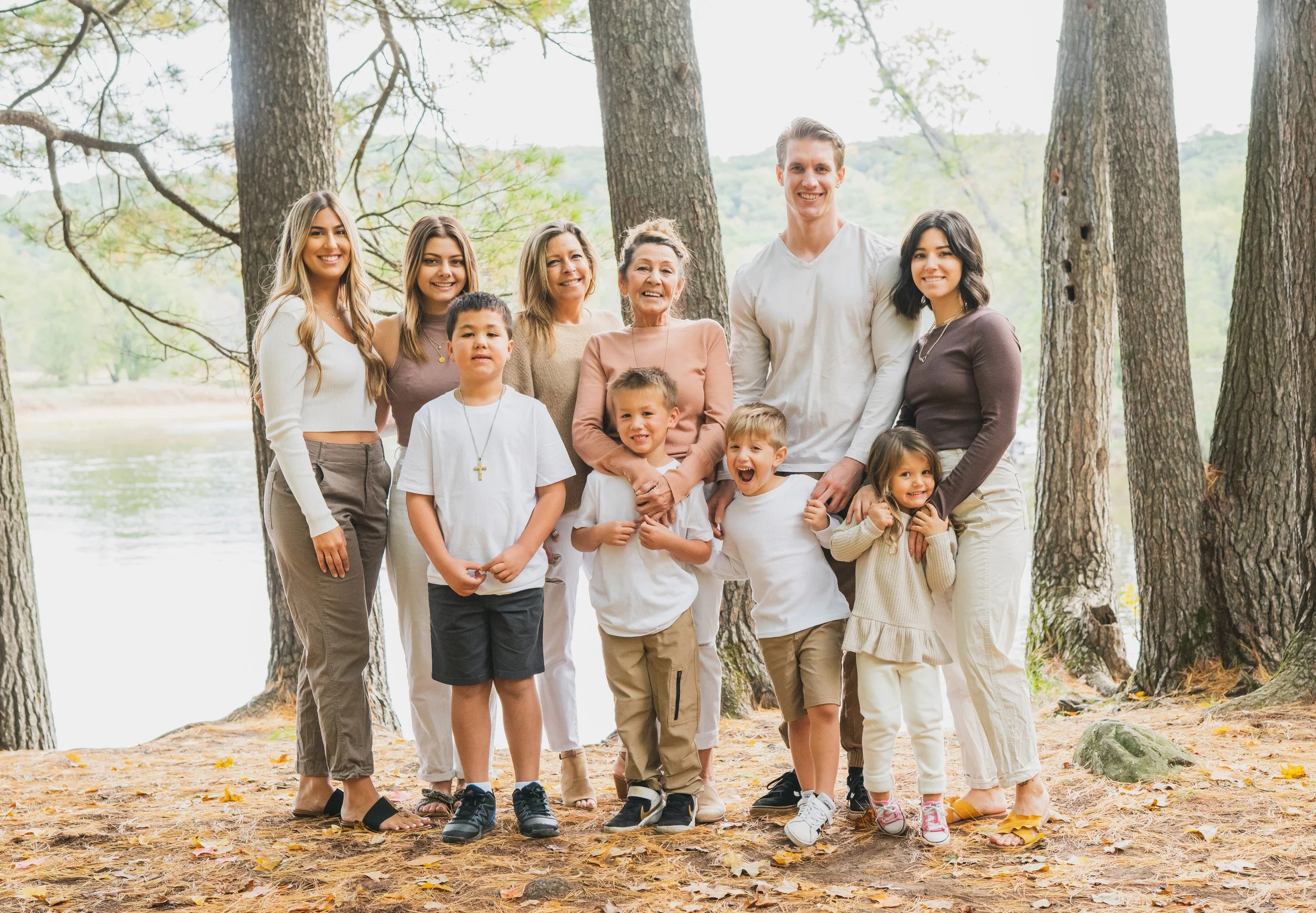 A multi-generational family of twelve posing together outdoors in a wooded area near a lake, smiling at the camera.