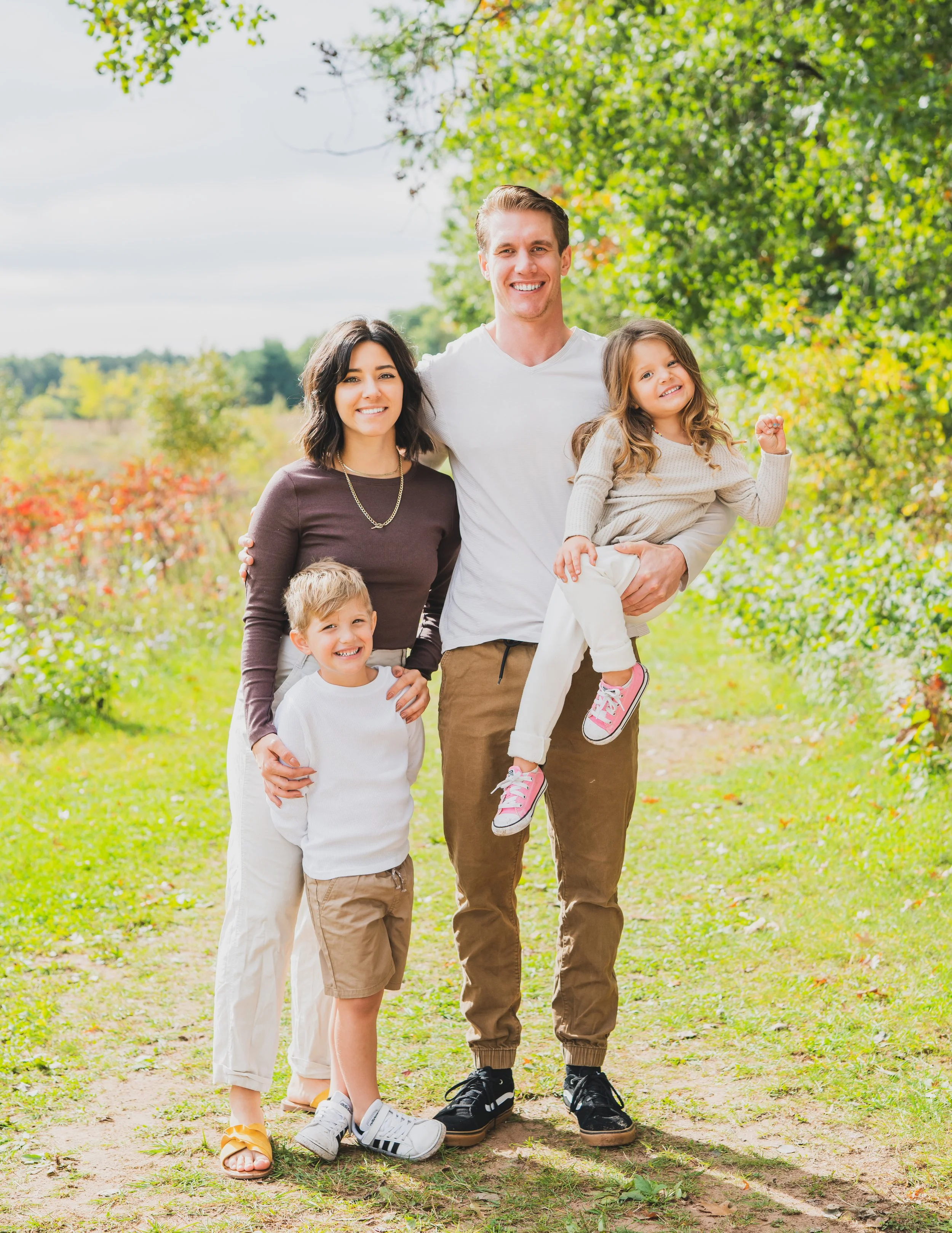 A happy family of five standing outdoors on a sunny day among trees and greenery, smiling at the camera.
