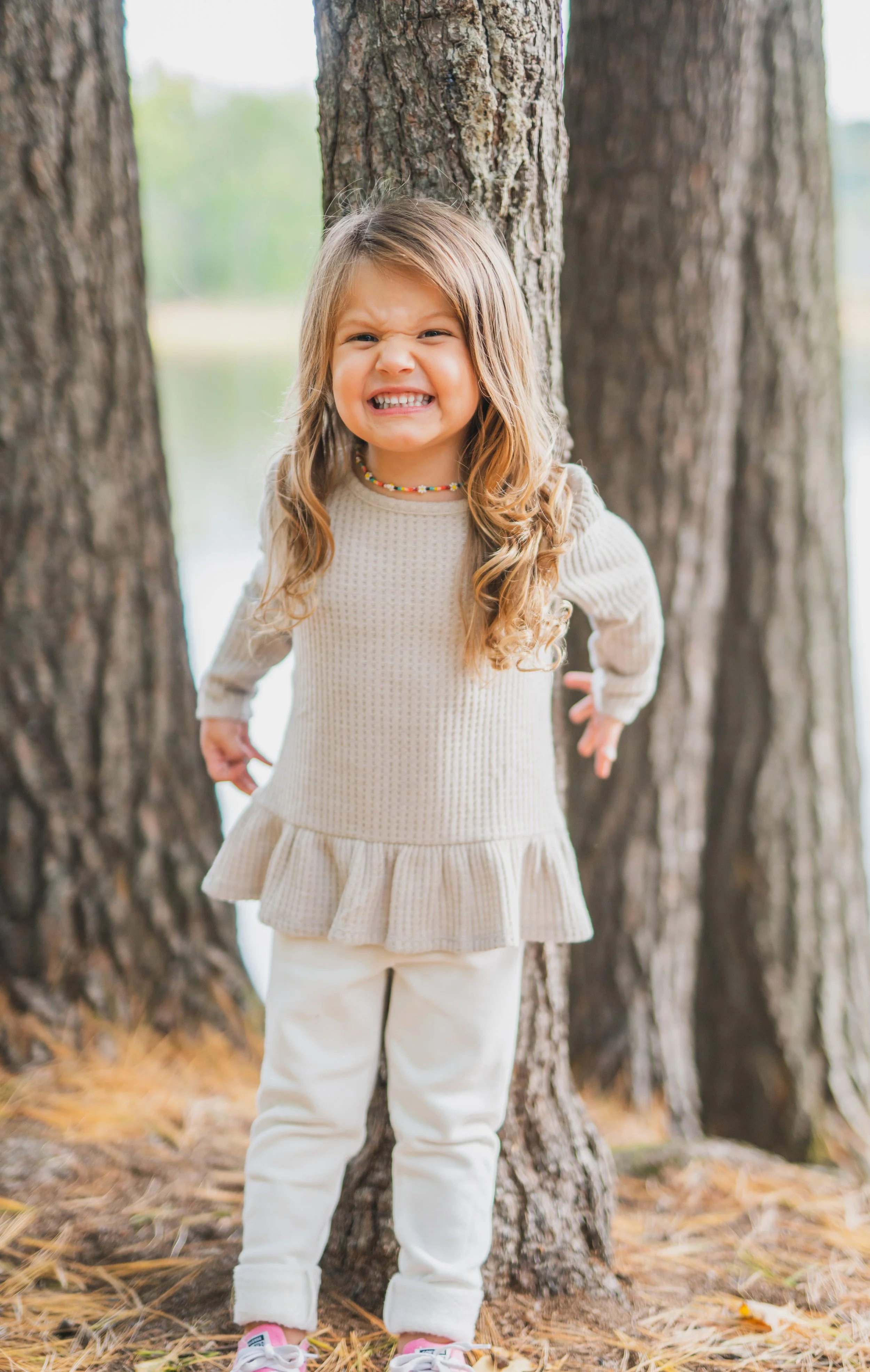 A young girl with long curly hair smiling outdoors among tall trees, wearing a beige sweater, white pants, pink sneakers, and a colorful beaded necklace.