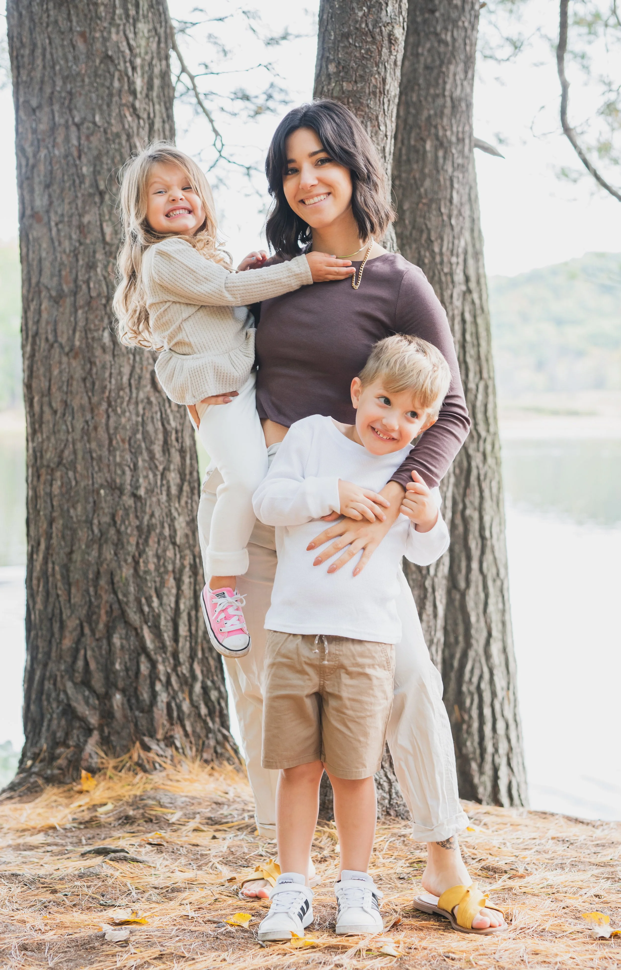 A woman with two children standing outdoors near trees and a body of water in the background, smiling at the camera.