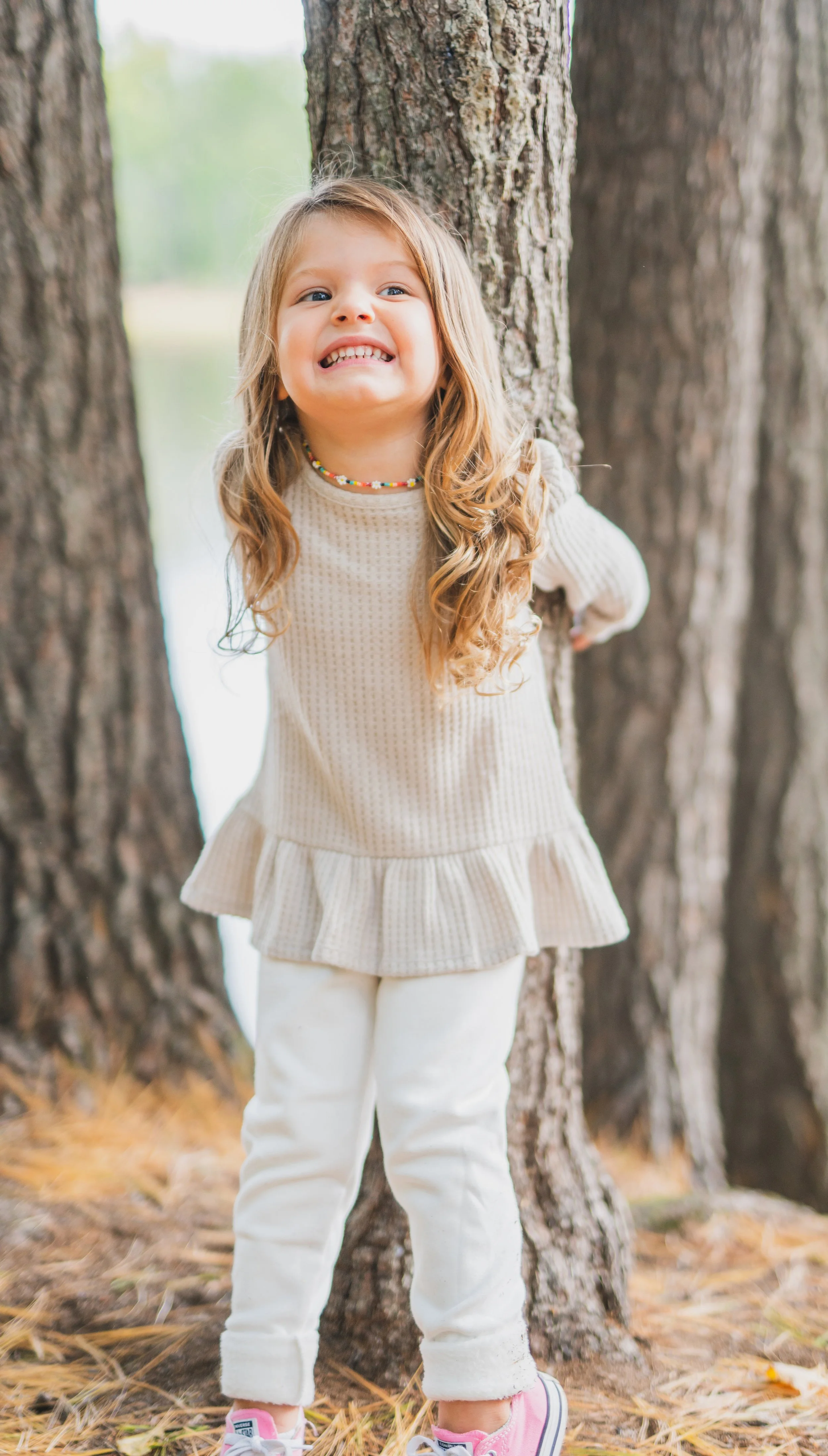 A young girl with long, curly blonde hair smiling playfully while standing among trees in a wooded area.