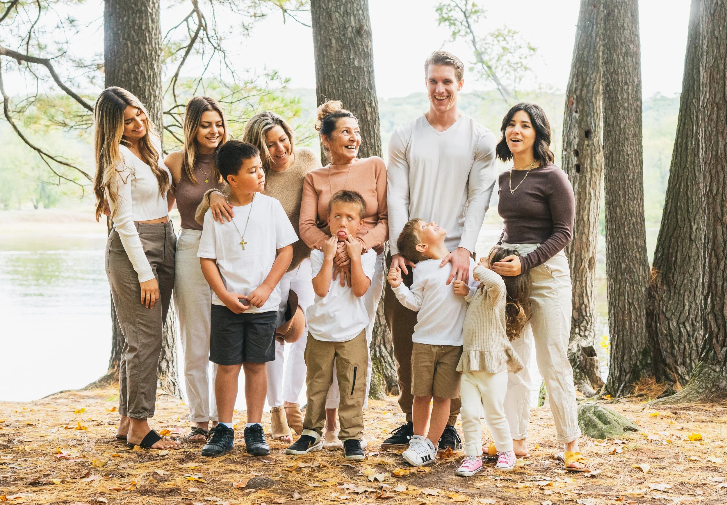 A large multigenerational family laughing and smiling outdoors in a park by a lake, surrounded by trees.