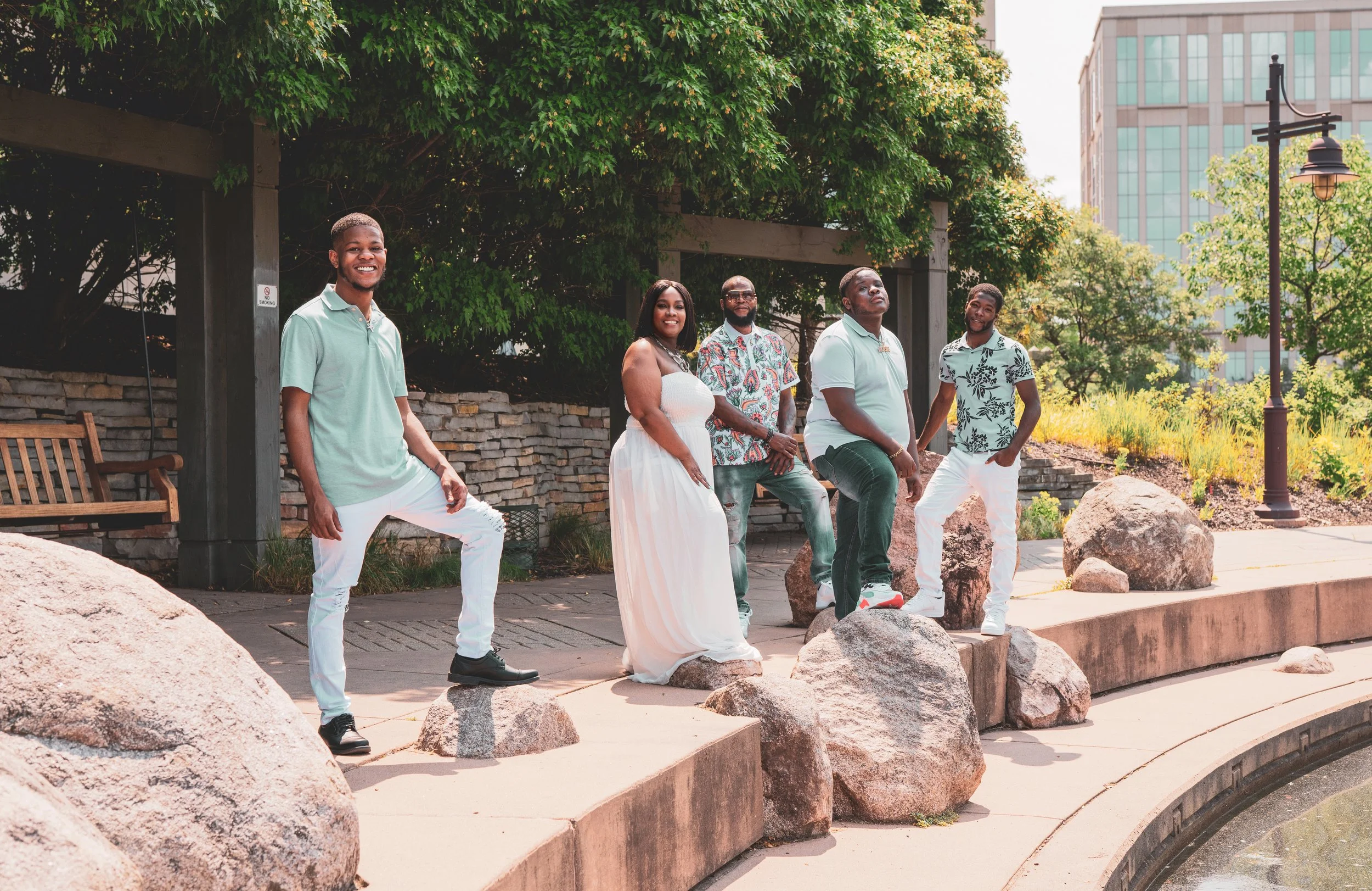 A group of six young adults standing and sitting on rocks near a pond in an outdoor park during daytime. They are smiling and dressed in casual summer clothing, with greenery and a city building in the background.