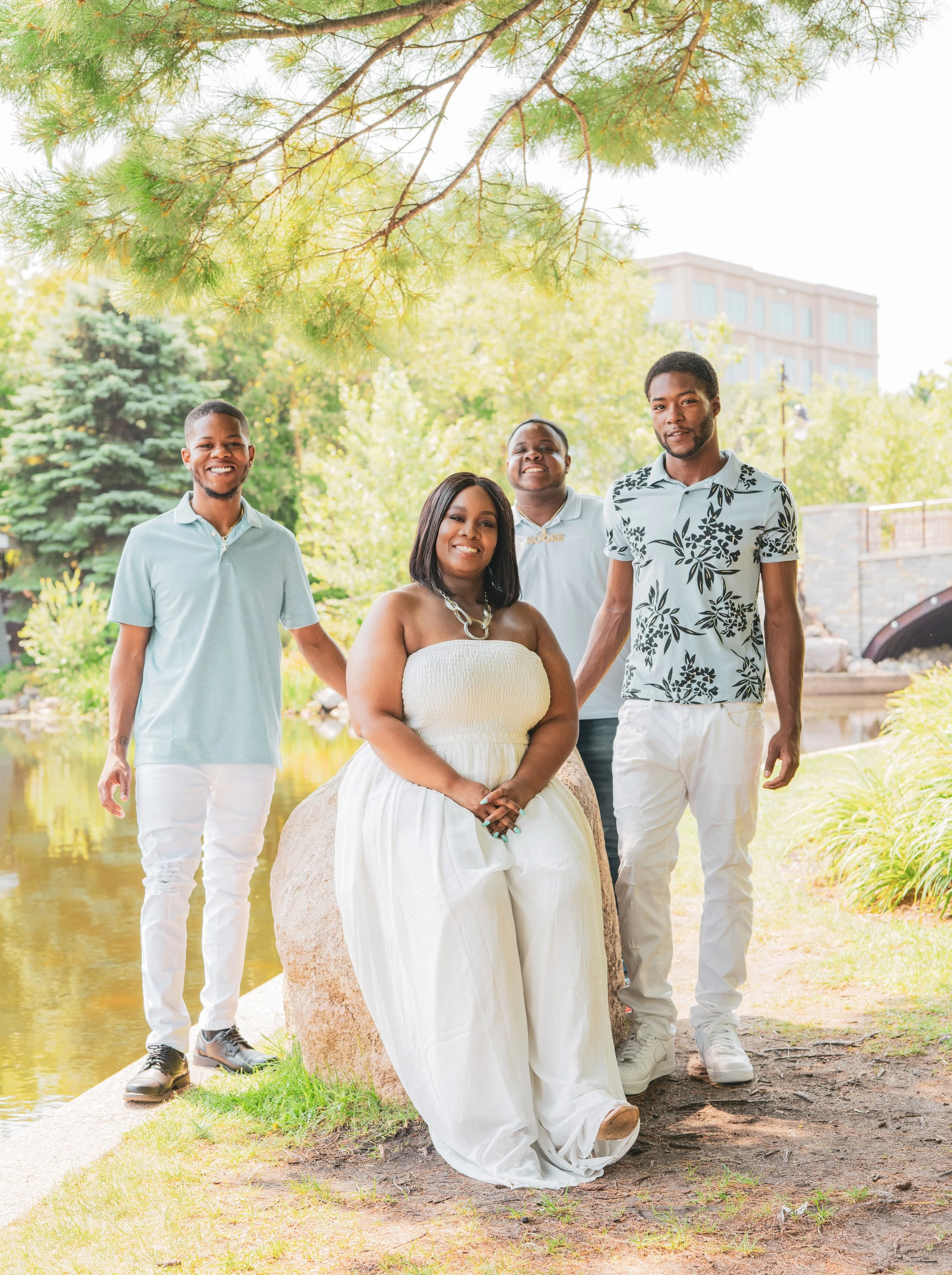 Group of four people, two men and two women, standing outdoors near a pond, smiling, with green trees and a building in the background.