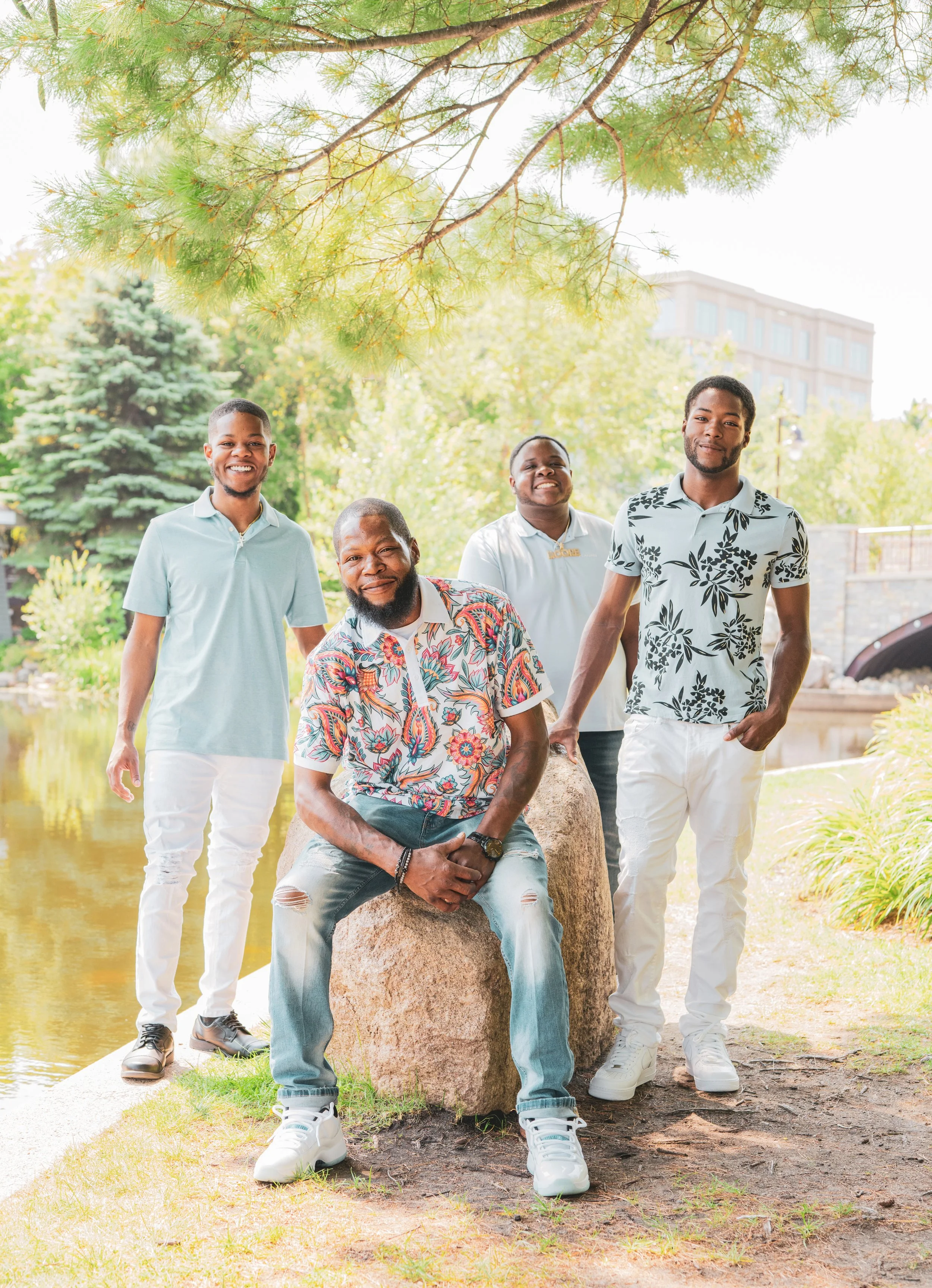 Four men standing and sitting near a pond in a park with trees and a building in the background, smiling for a photo.