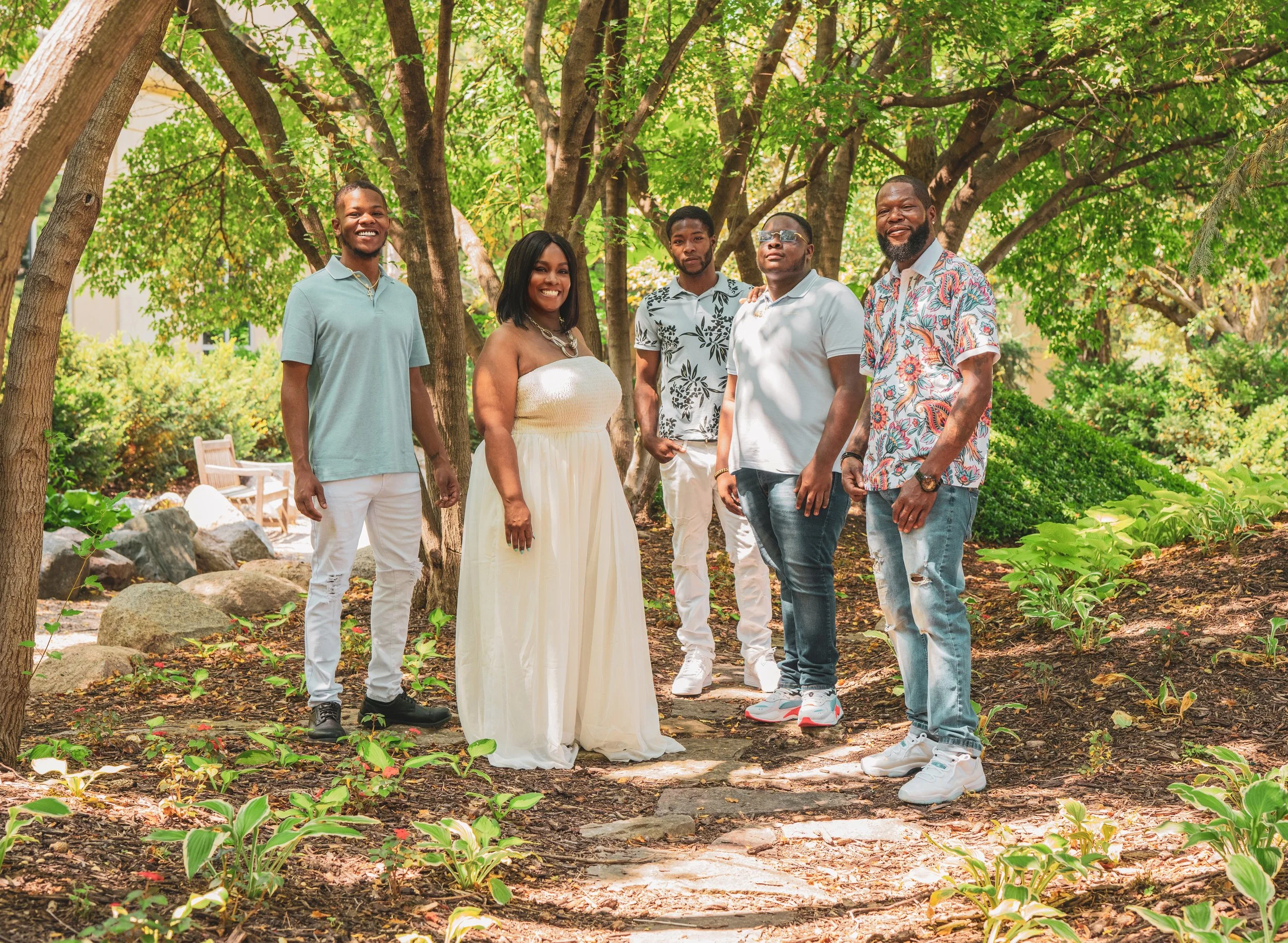 A group of six people standing on a garden path surrounded by greenery and trees, smiling at the camera.