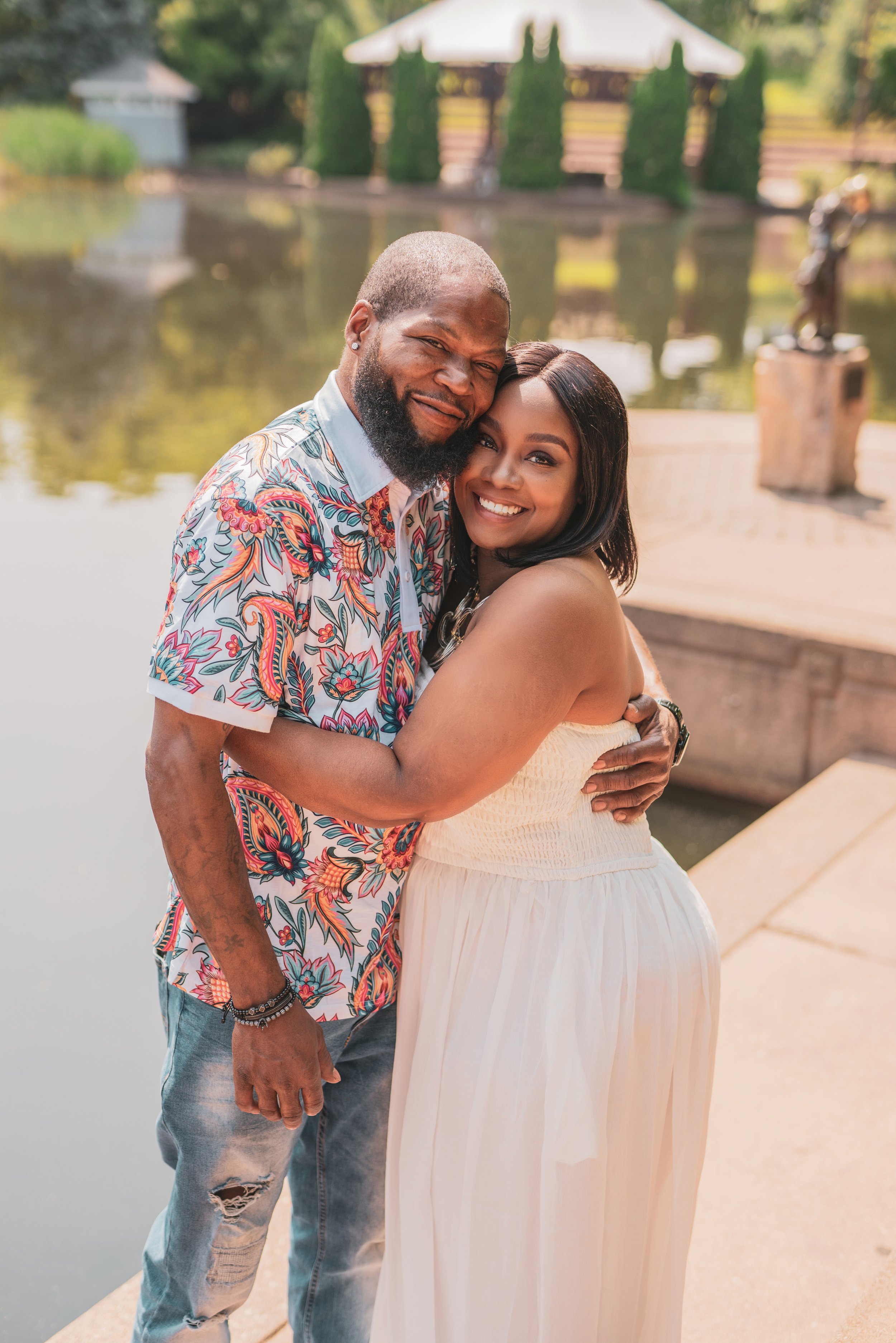 A smiling couple hugging by a pond with trees and a gazebo in the background.