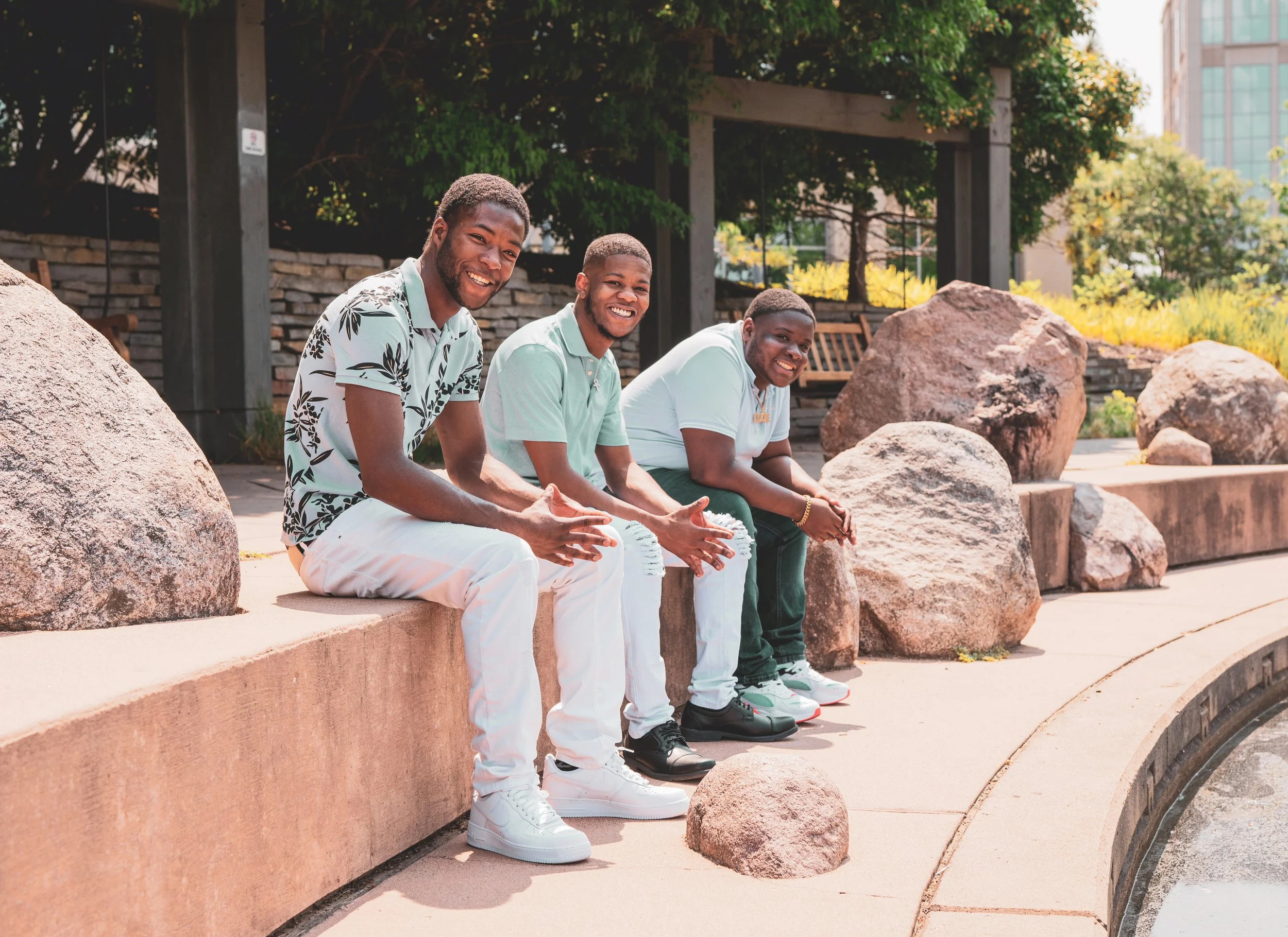 Three young men sitting on a ledge outdoors, smiling and looking at the camera, with large rocks, trees, and a building in the background.