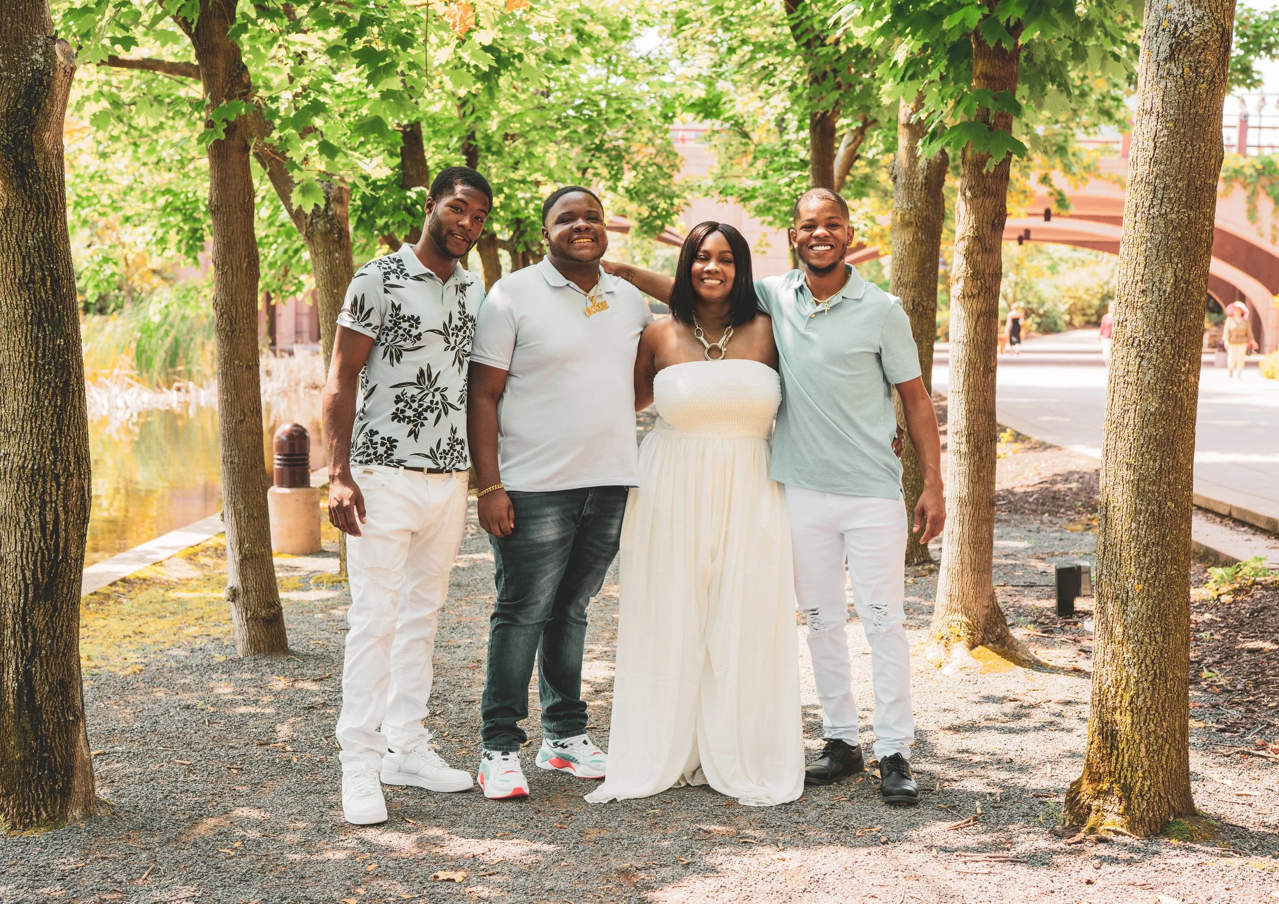 Group of four friends standing outdoors under trees, smiling, with a pond and pathway in the background.