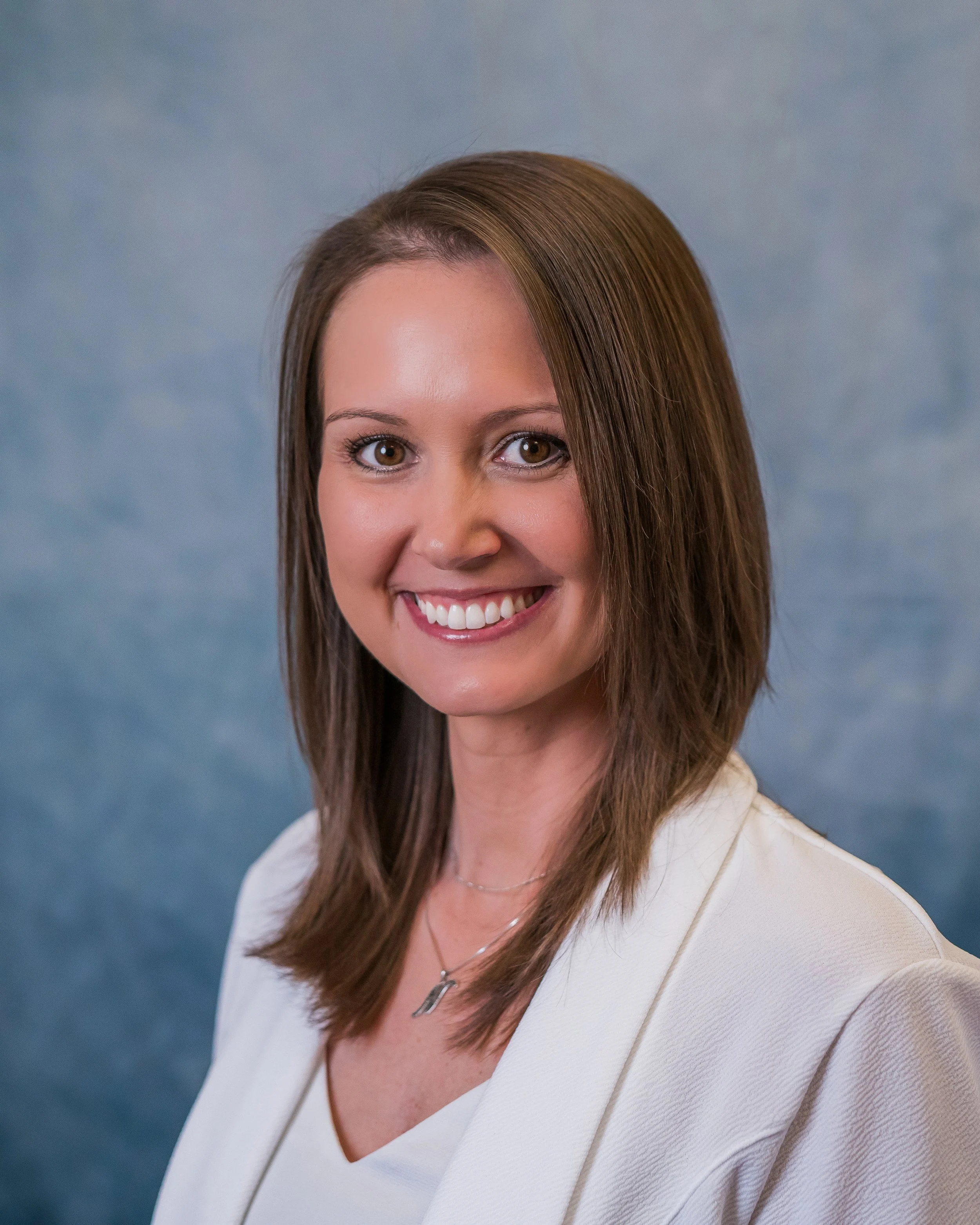 A smiling woman with straight, shoulder-length brown hair, wearing a white blazer and a delicate necklace, against a blue mottled background.