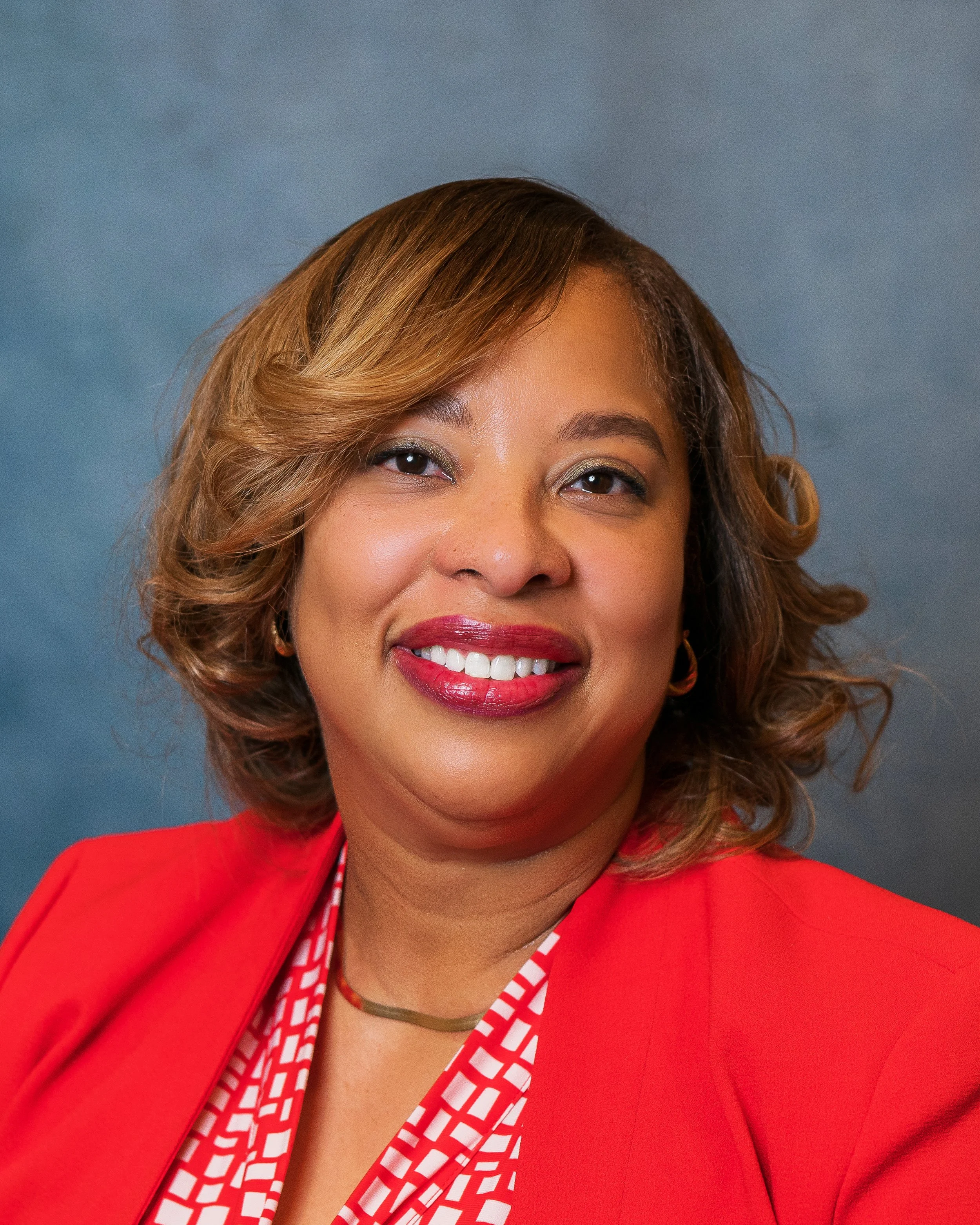 A woman with shoulder-length wavy hair, wearing a red blazer and smiling, posed against a blue background.