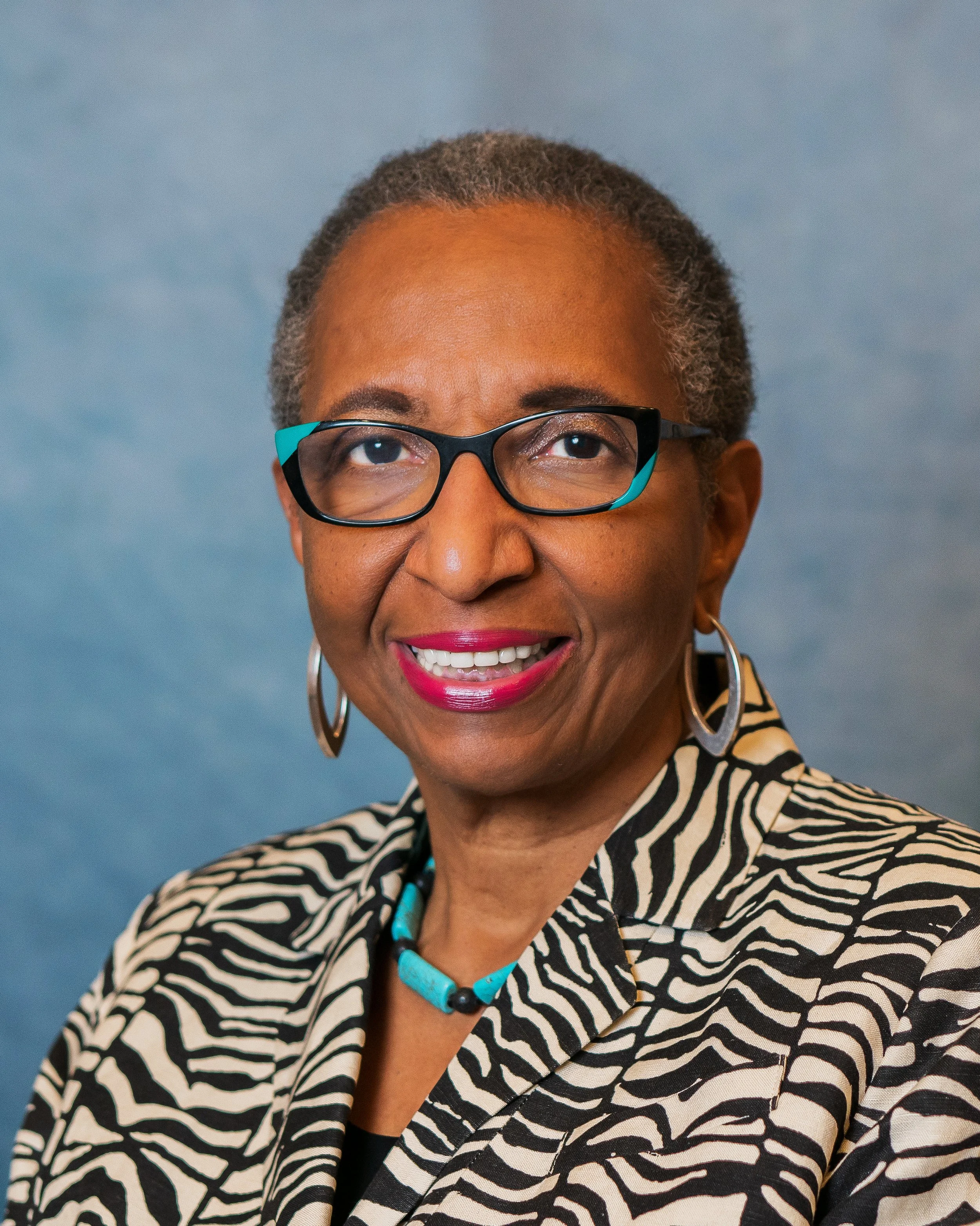 A portrait of an African American woman with short gray hair, wearing black and teal glasses, hoop earrings, a turquoise necklace, a black top, and a zebra print blazer, smiling against a light blue background.