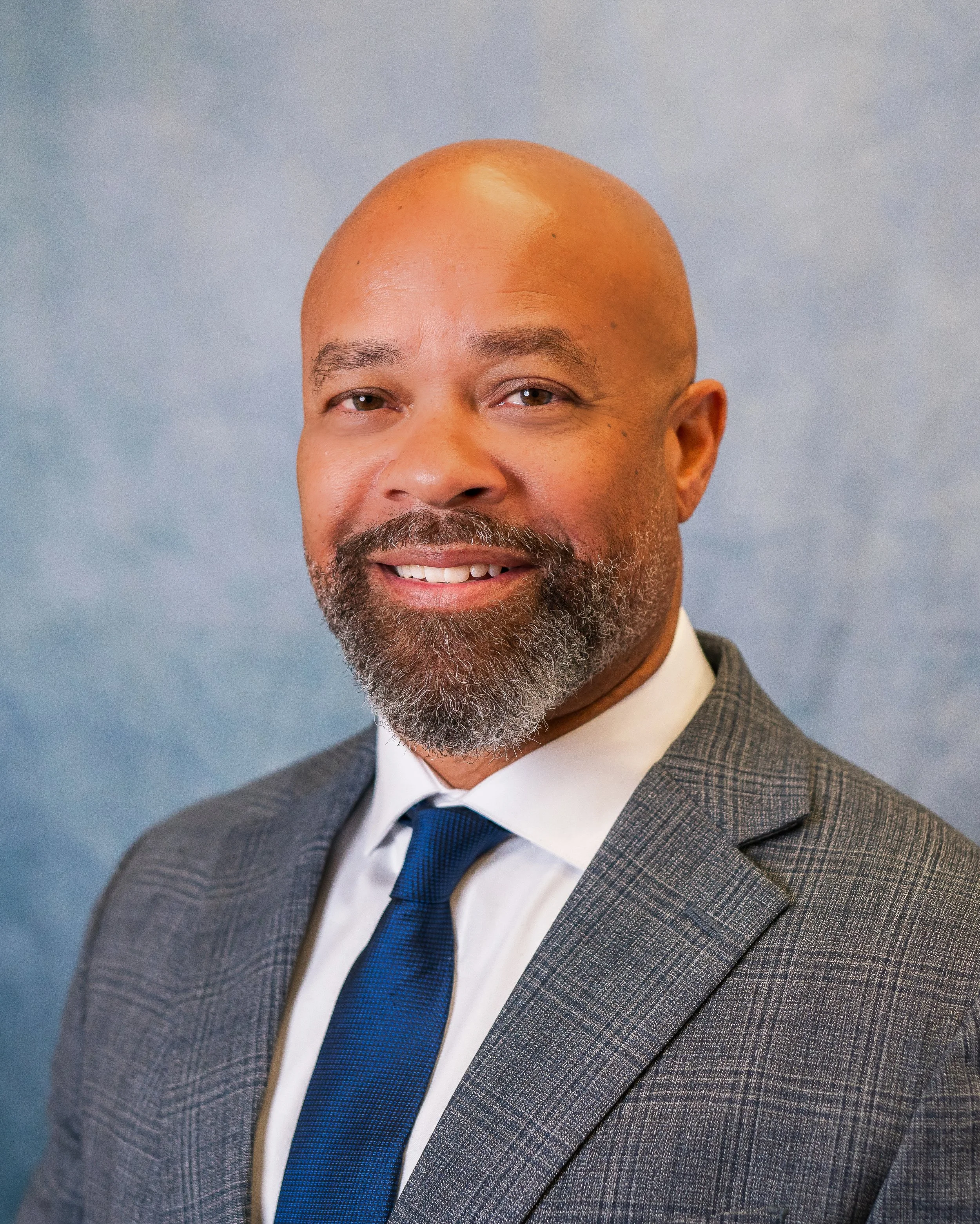 Headshot of a smiling, middle-aged African American man with a bald head and gray beard, wearing a gray checked suit, white shirt, and blue tie, against a blue background.