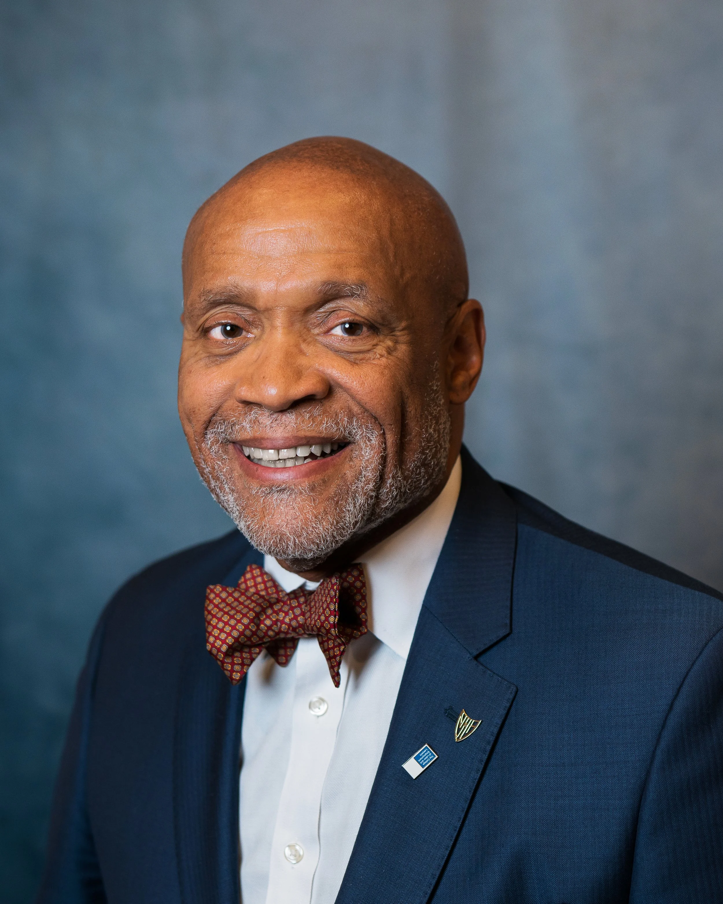A smiling African American man in a dark suit with a white shirt, red patterned bow tie, and two pins on his lapel, in front of a blue background.