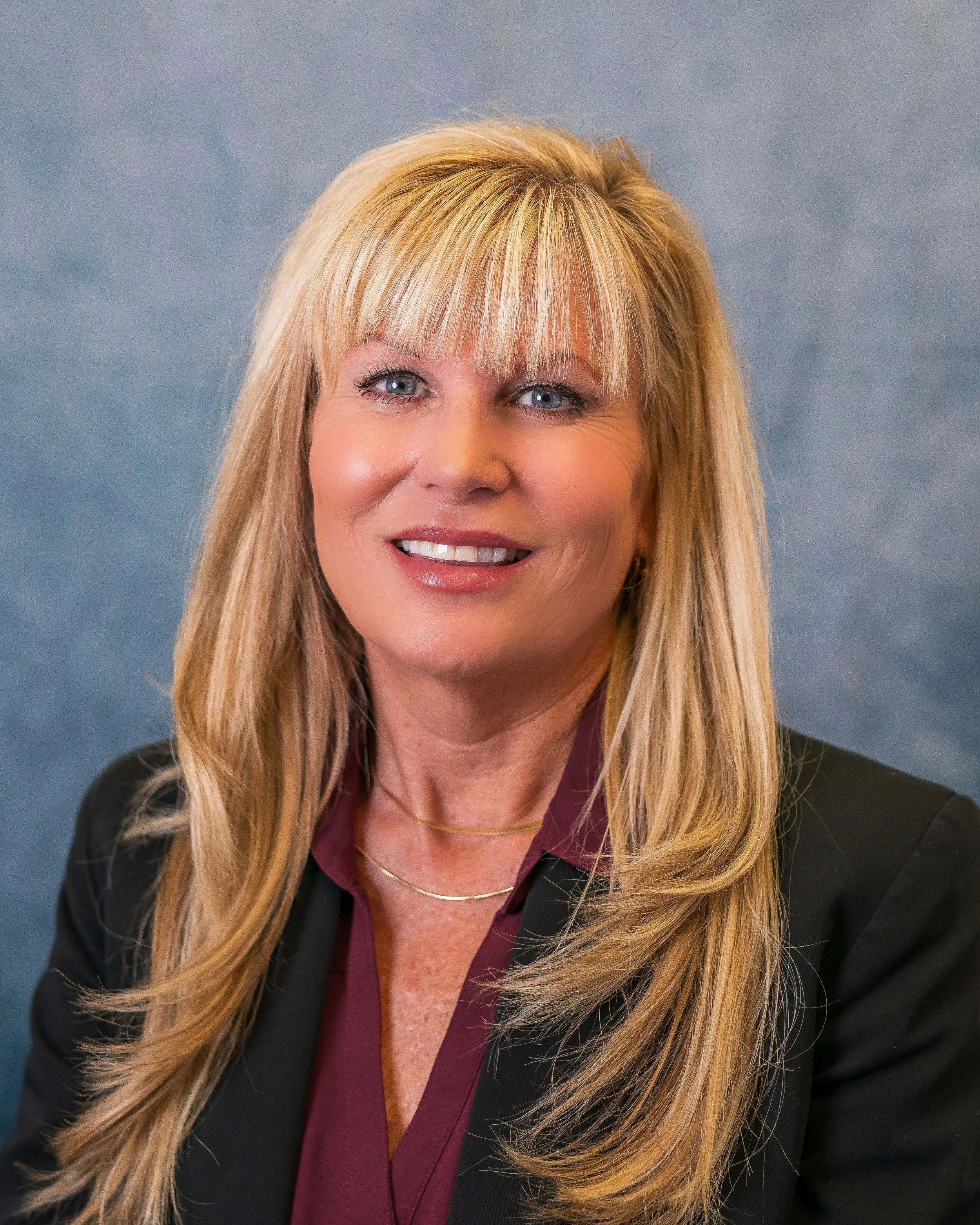 A woman with blonde hair and blue eyes smiling, wearing a black blazer and a maroon blouse, posed against a gray textured background.