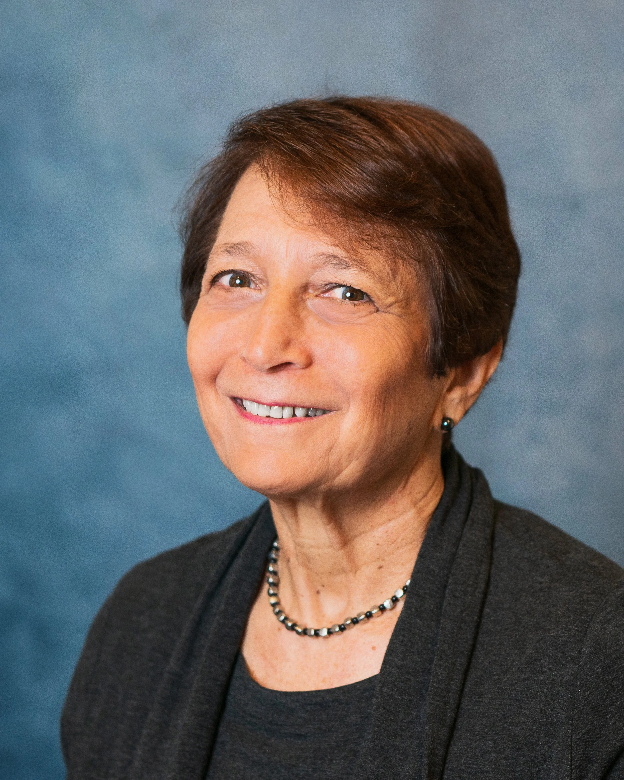 A portrait of an older woman with short brown hair, smiling, wearing a black blazer, a beaded necklace, and matching earrings, against a blue background.