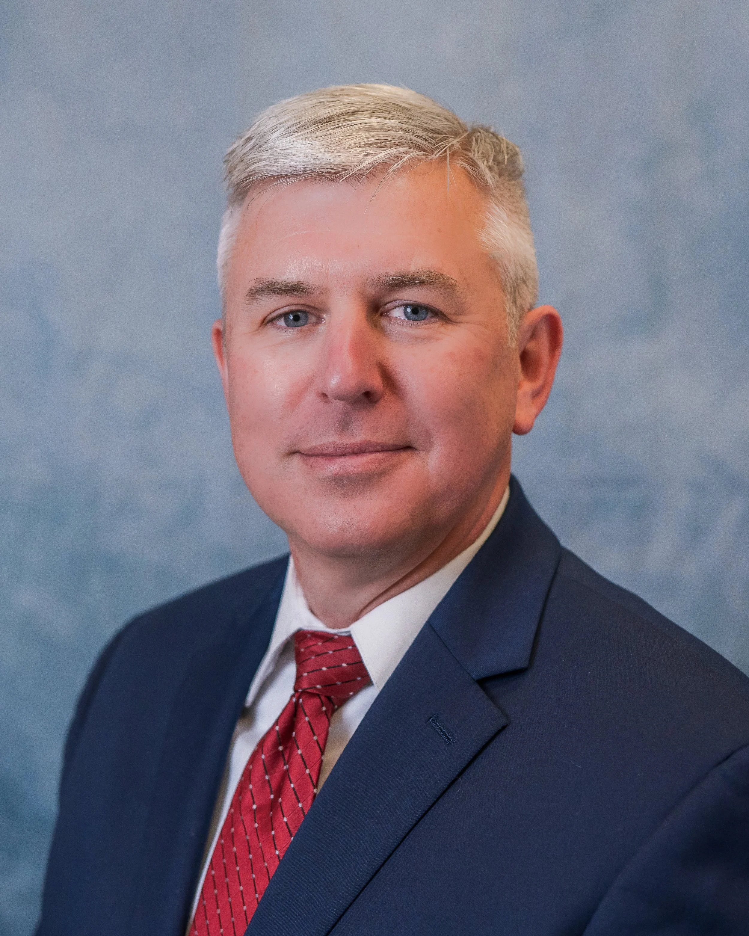 A professional man with blond hair and blue eyes, wearing a navy suit, white shirt, and red tie, posing in front of a plain gray background.