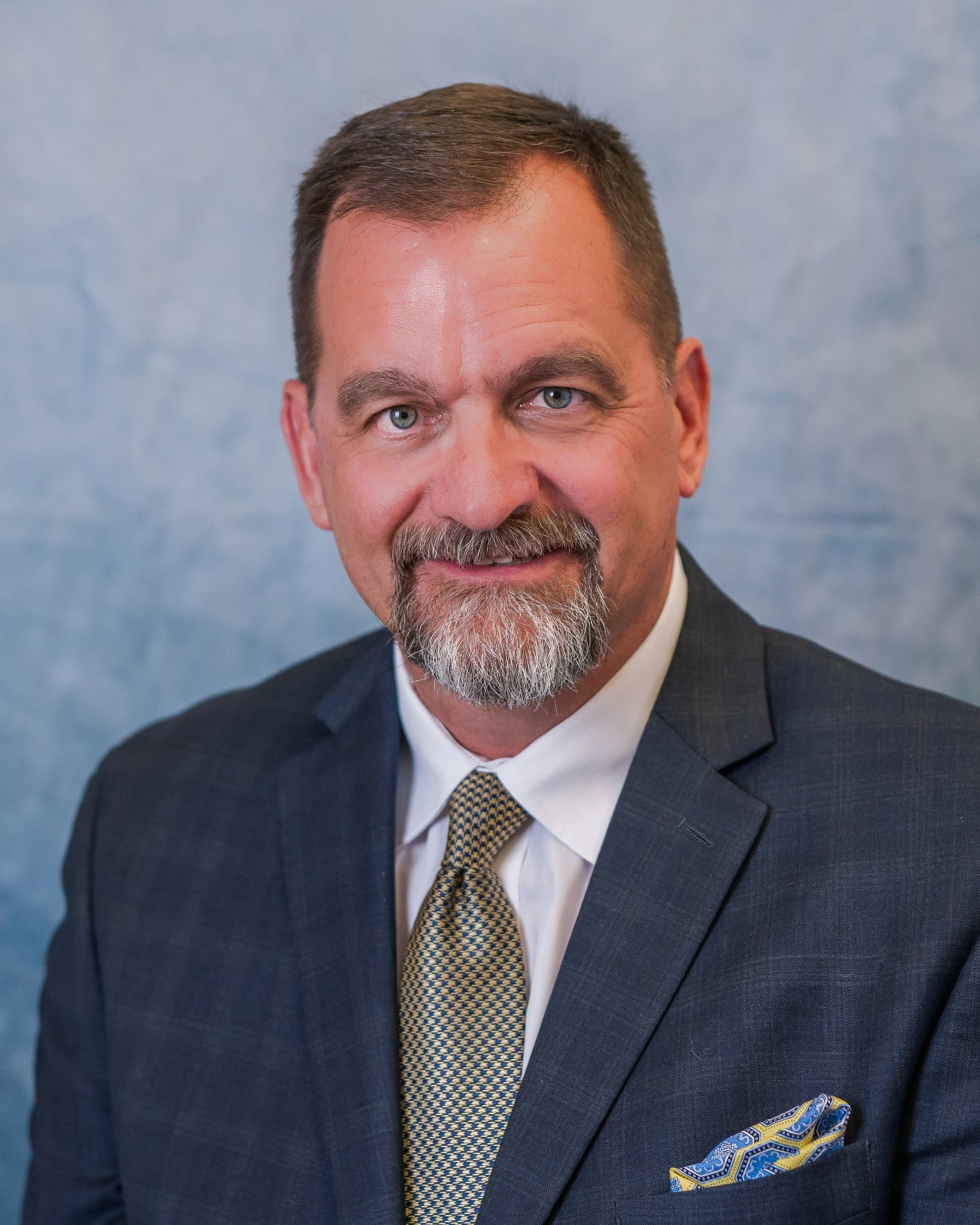 A professional man in a suit, white shirt, and patterned tie, with a matching pocket square, smiling against a blue background.