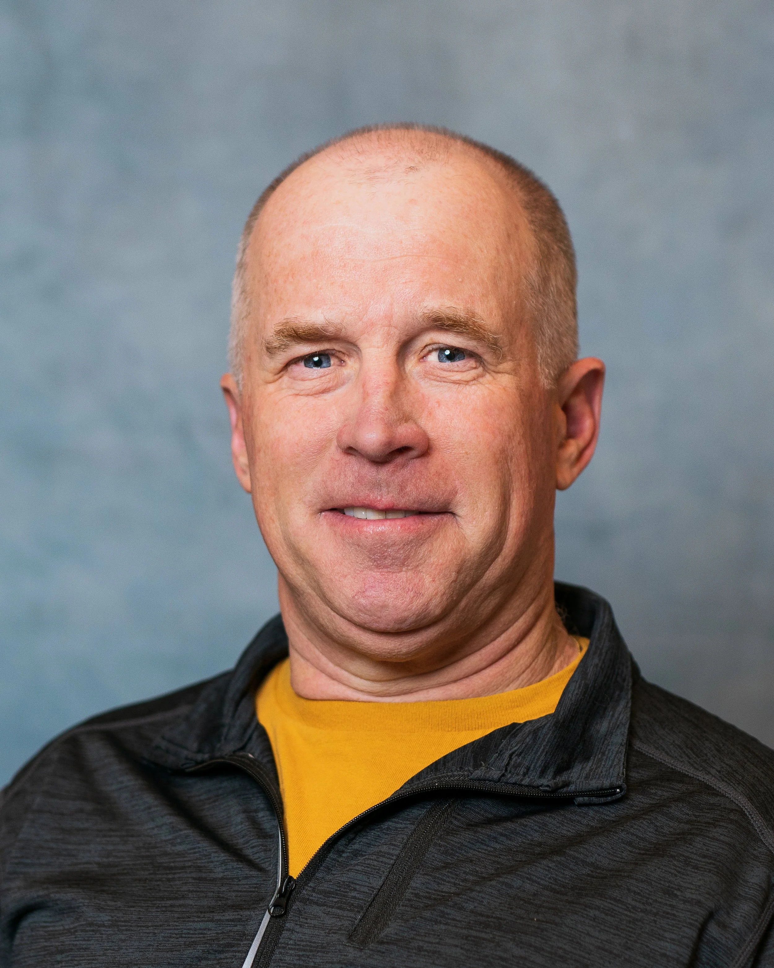 Close-up of a smiling middle-aged man with a shaved head, light blue eyes, wearing a black zip-up jacket over a yellow shirt, against a neutral background.