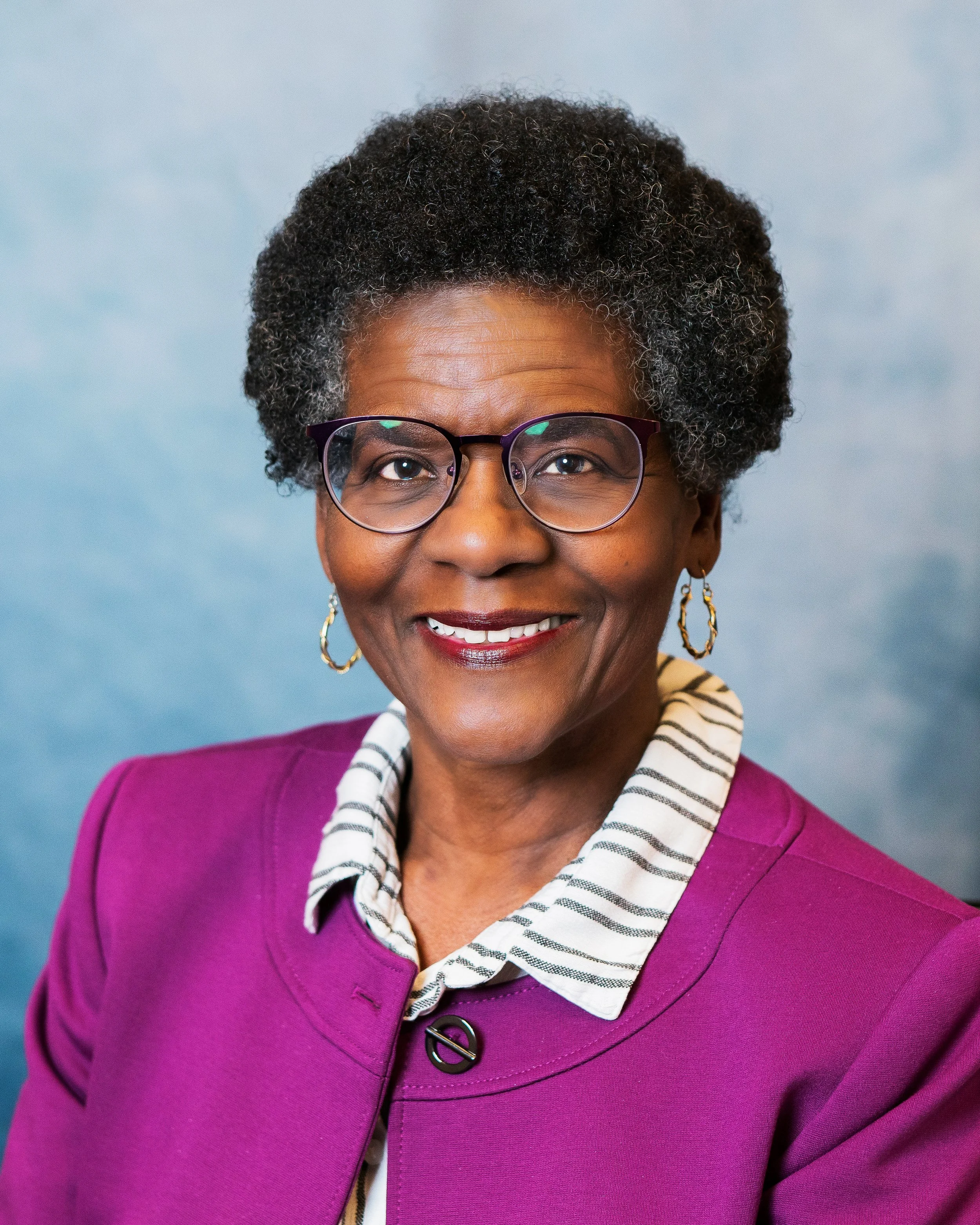 A portrait of an older African American woman with short, curly gray hair, wearing glasses, gold hoop earrings, a magenta blazer, and a striped collared shirt, smiling against a blue background.