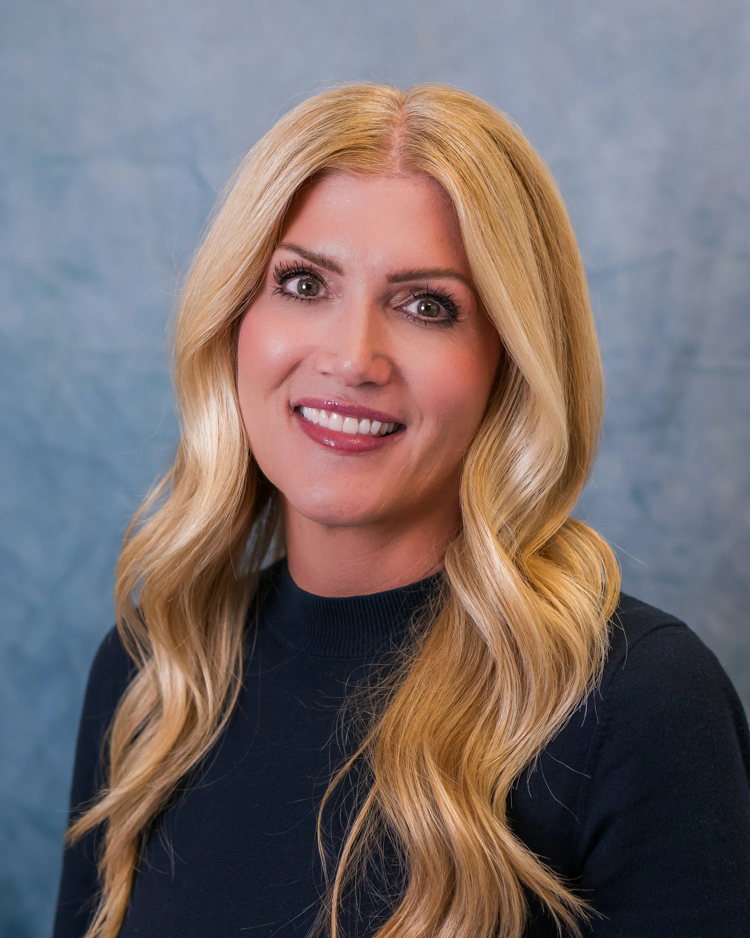 A woman with long, wavy blonde hair smiling at the camera, wearing a black top, against a blue textured background.