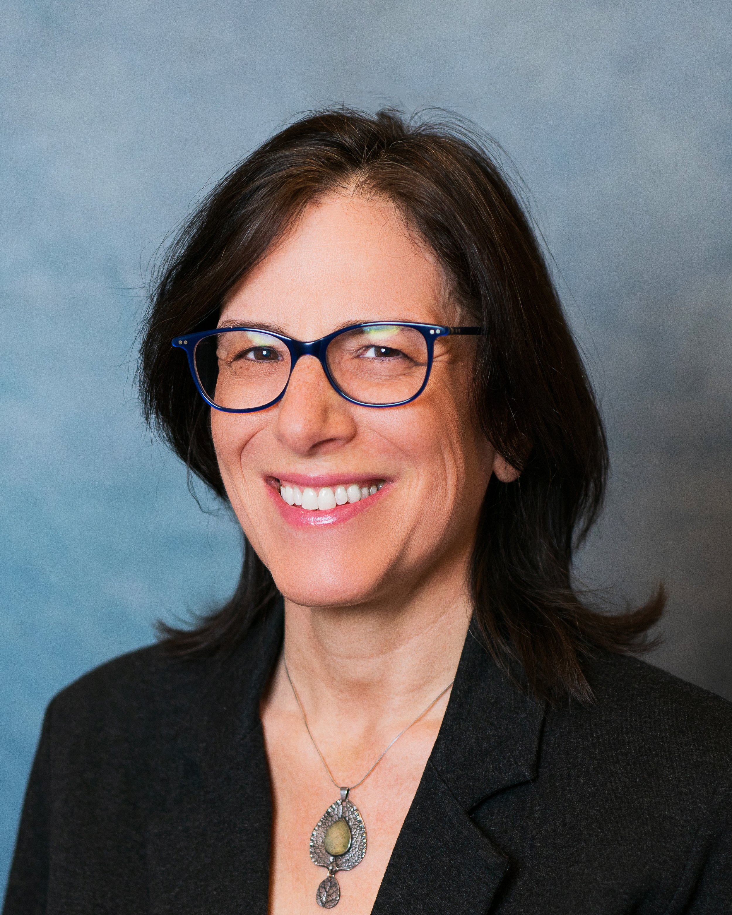 Portrait of a woman with dark hair, wearing glasses and a black blazer, smiling at the camera against a blue background.