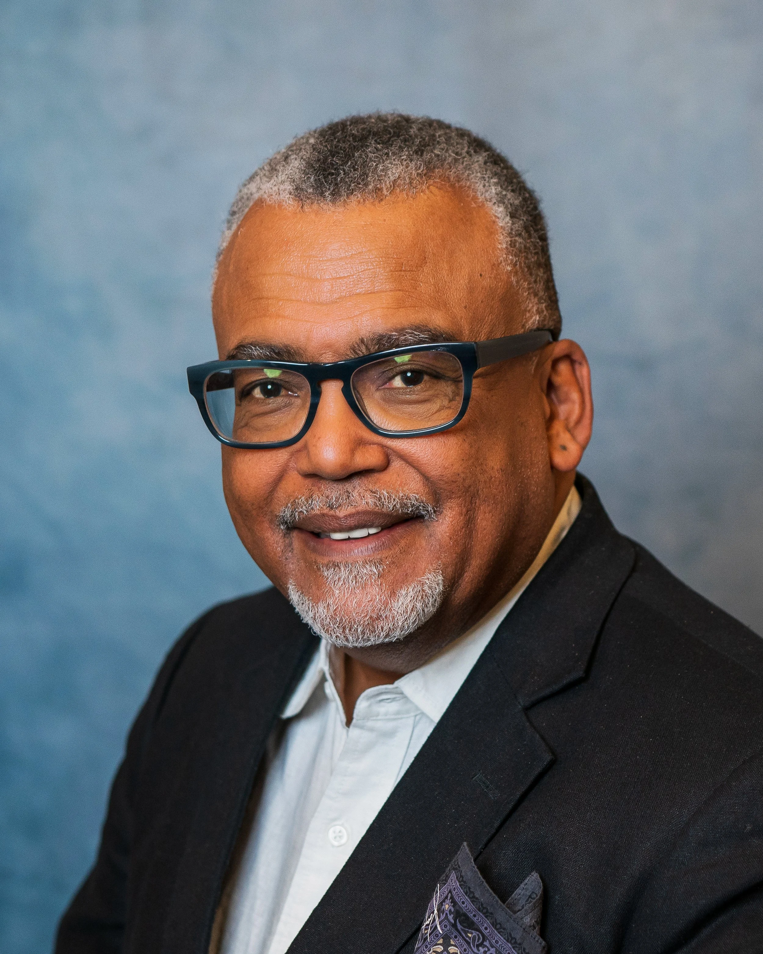 A smiling middle-aged man with gray hair and a beard, wearing glasses, a black blazer, and a white shirt, posed against a blue background.