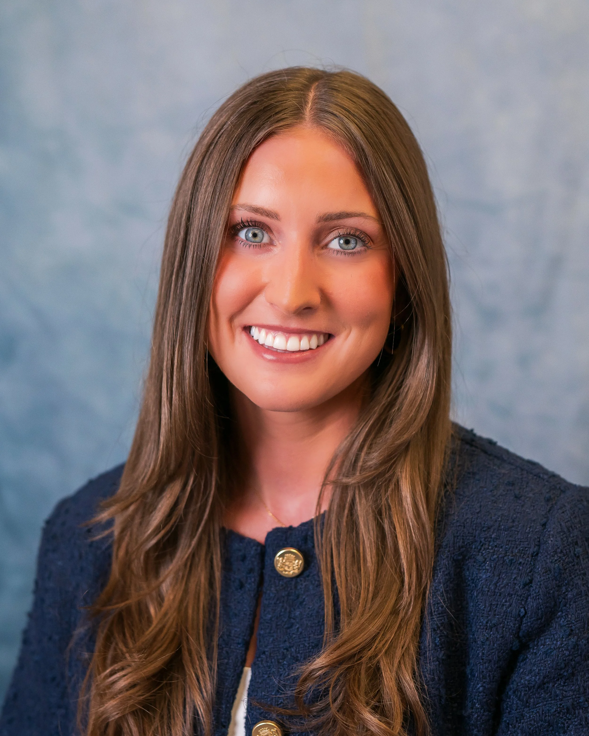 A woman with long light brown hair and blue eyes smiling, wearing a navy blazer with gold buttons, posing against a light blue textured background.