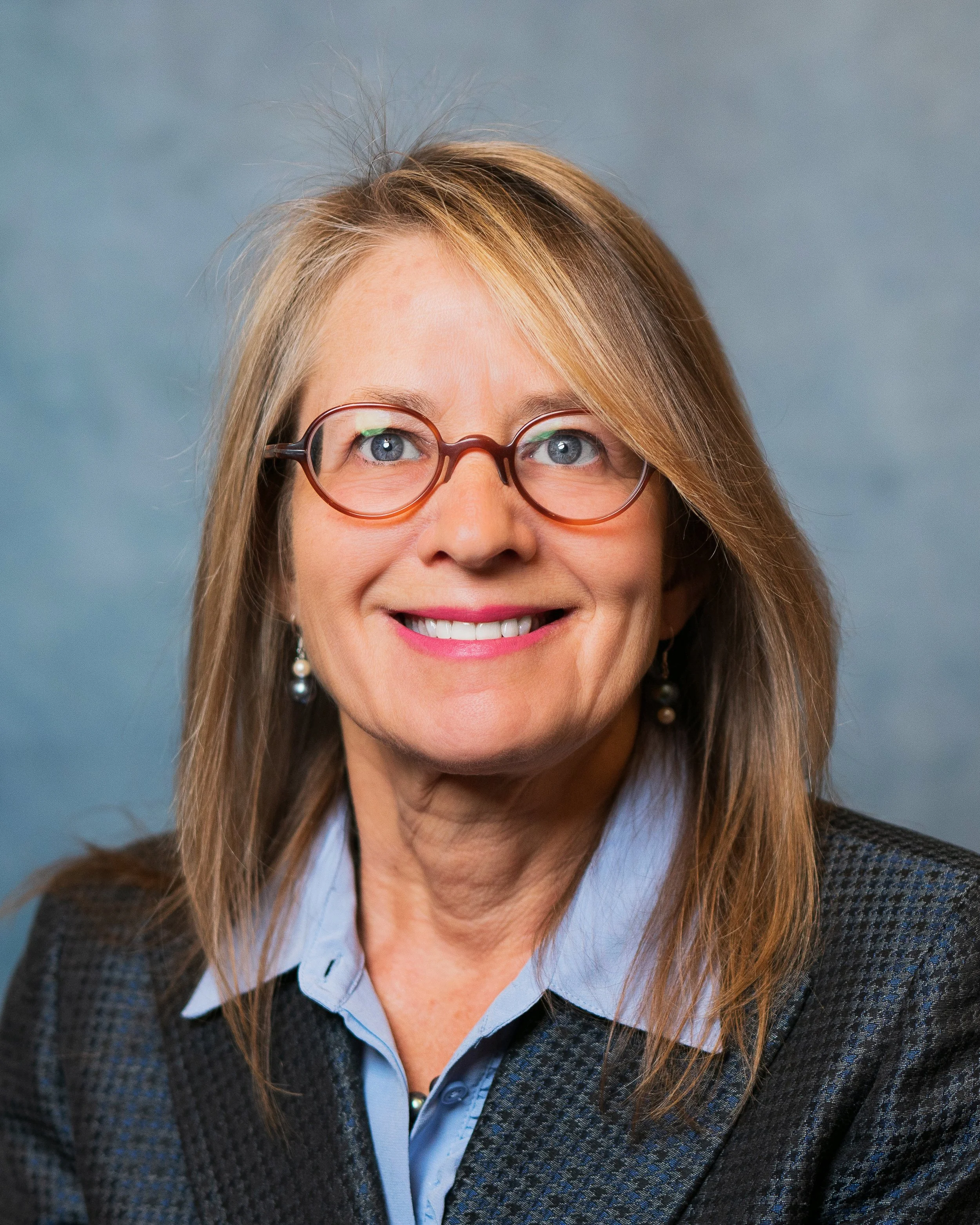 A woman with light brown hair, wearing glasses, a blue collared shirt, and a dark blazer, smiling against a gray-blue background.